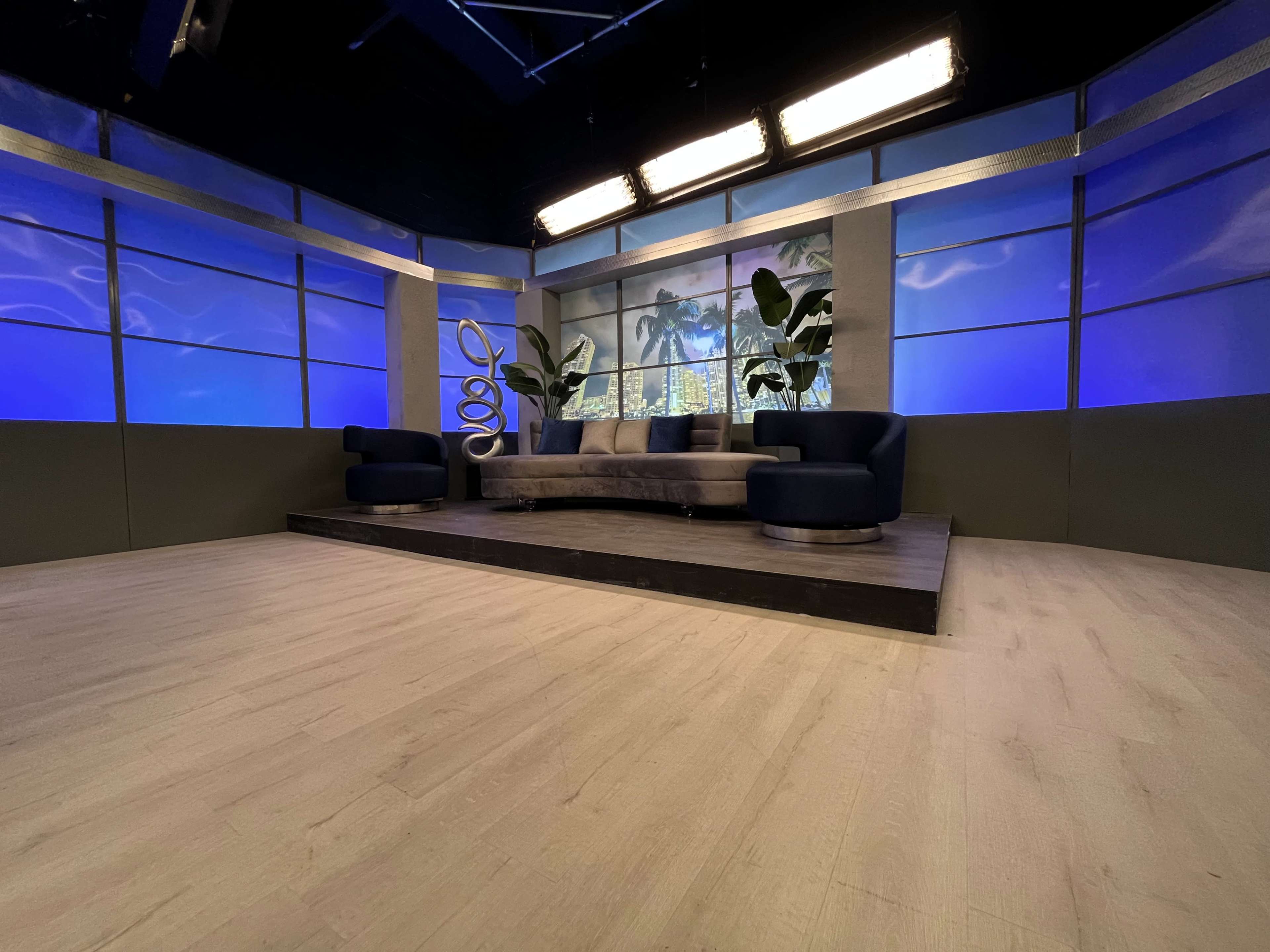 A television studio set with a curved sofa, blue accents, and large windows displaying palm trees in the background.
