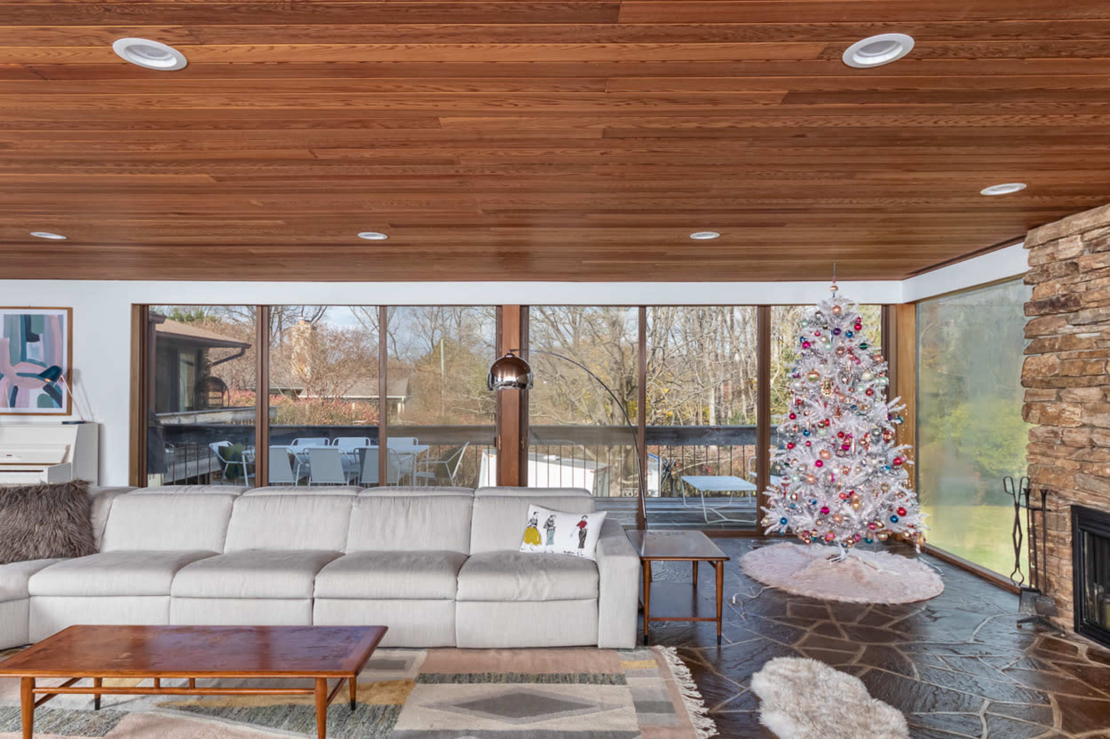 A modern living room features a white Christmas tree decorated with colorful ornaments, a large sectional sofa, and a stone fireplace, with large windows providing a view of the outdoors.
