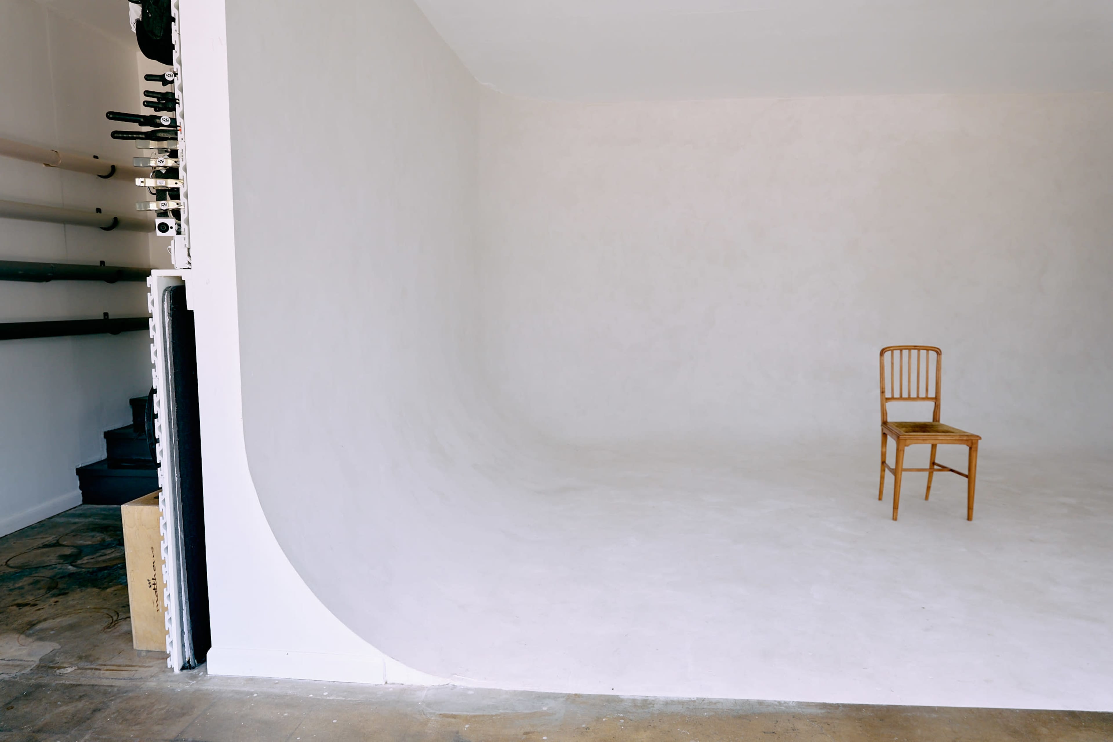 The image shows a minimalist studio space with a smooth, curved white wall and a single wooden chair positioned in the center.