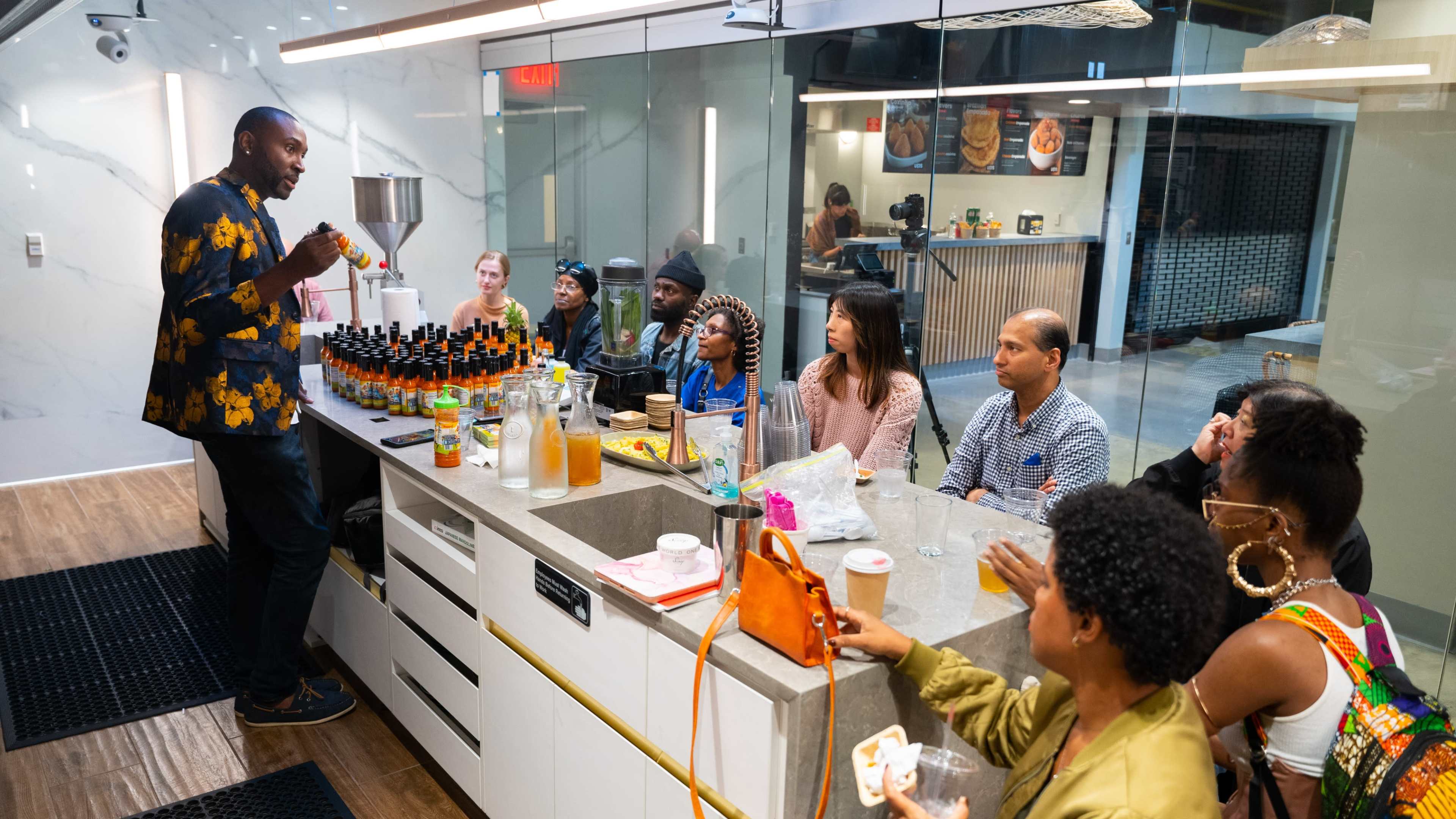 A speaker presents to a group seated around a countertop in a modern kitchen setting, surrounded by various beverages and ingredients.