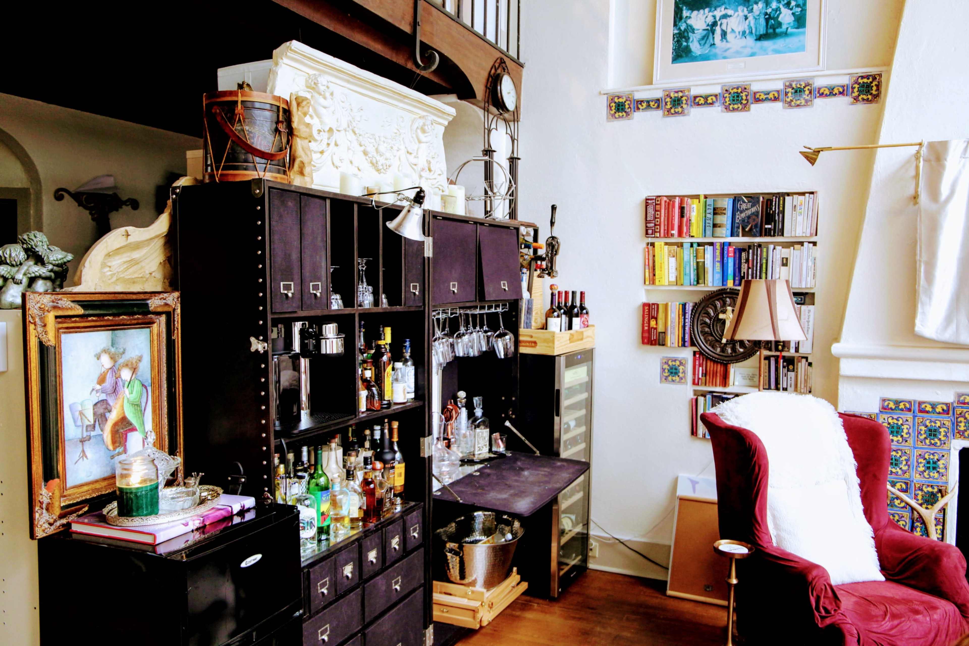A home bar area with a wooden cabinet displaying various bottles, glassware, and a framed painting, alongside a small bookshelf and a cozy armchair.