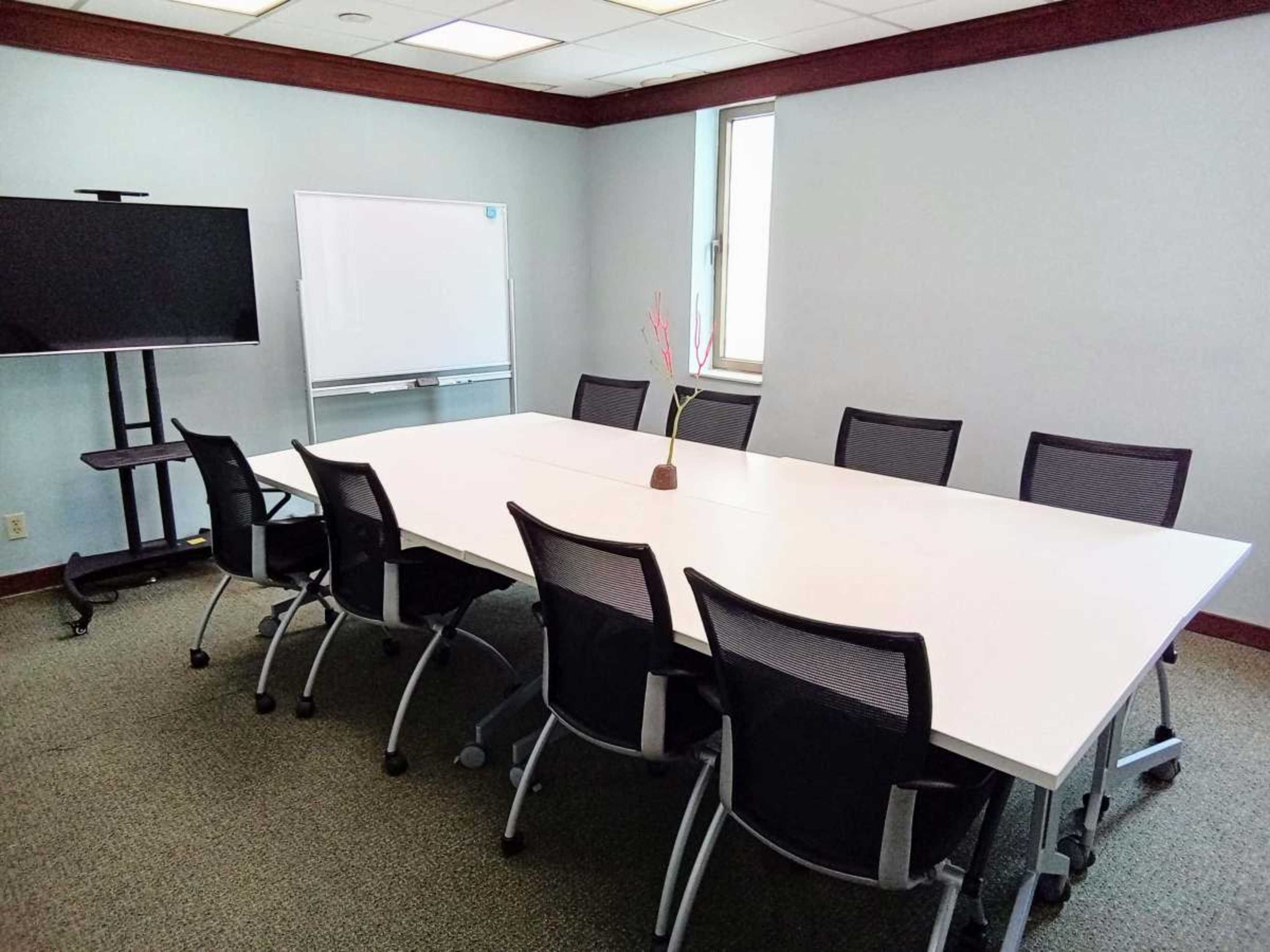 A conference room features a large white table surrounded by eight black chairs, with a television on a stand and a whiteboard in the background.