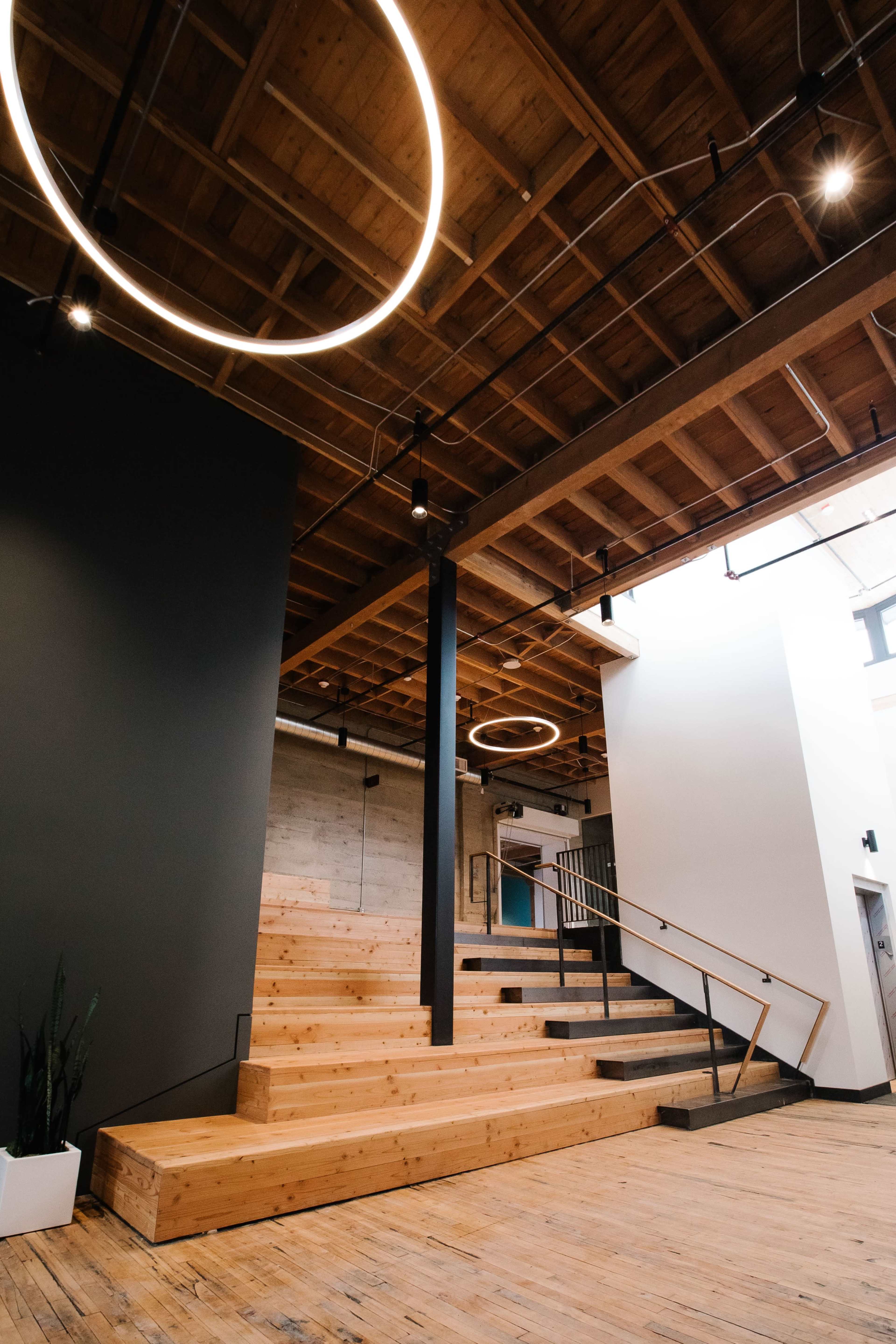 The image shows a modern interior space with wooden steps leading up to a platform, sleek railings, and circular light fixtures hanging from the ceiling.