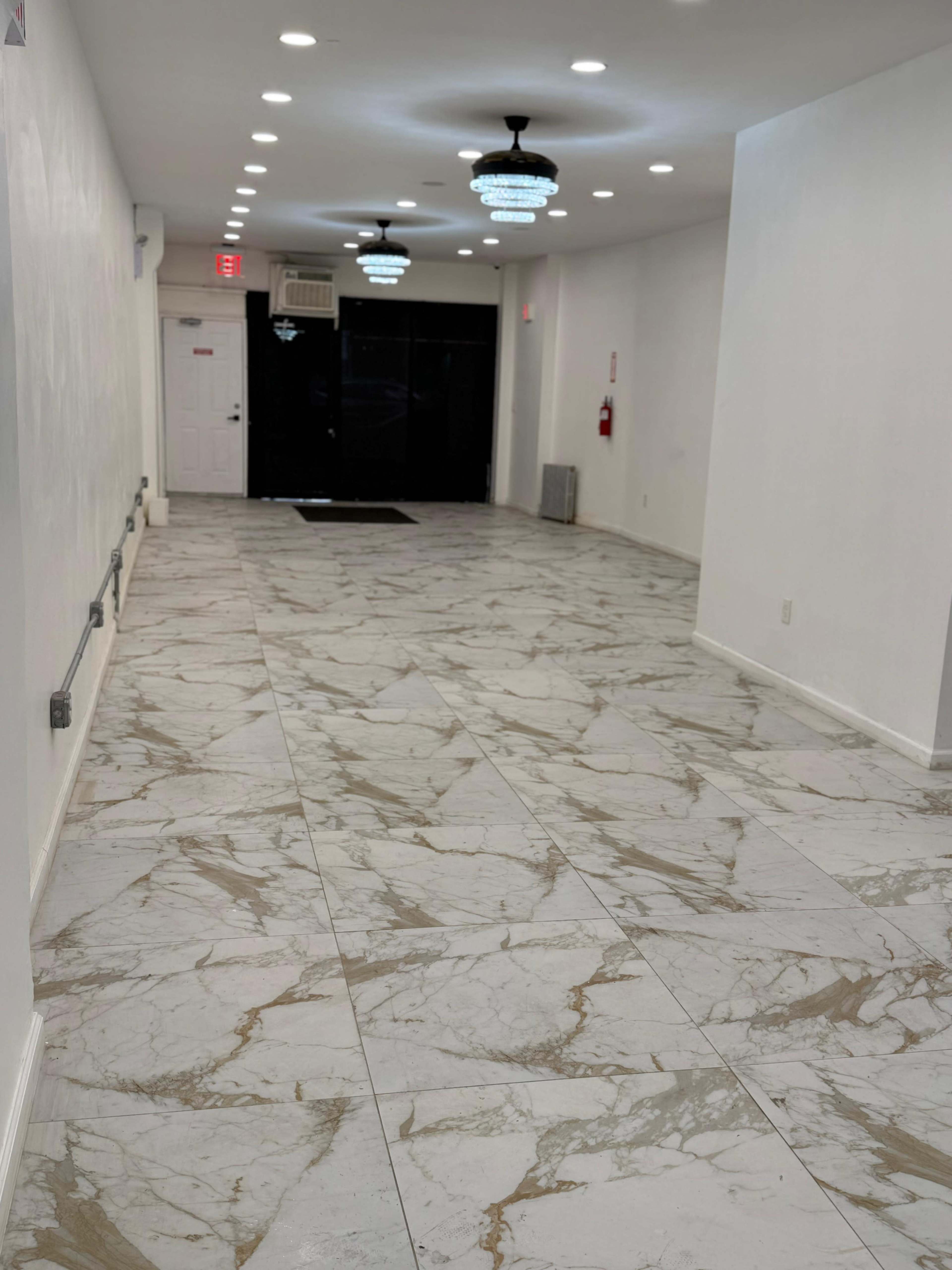 The image shows a long, white hallway with marble-patterned tile flooring and decorative ceiling lights, leading to a dark entrance at the far end.