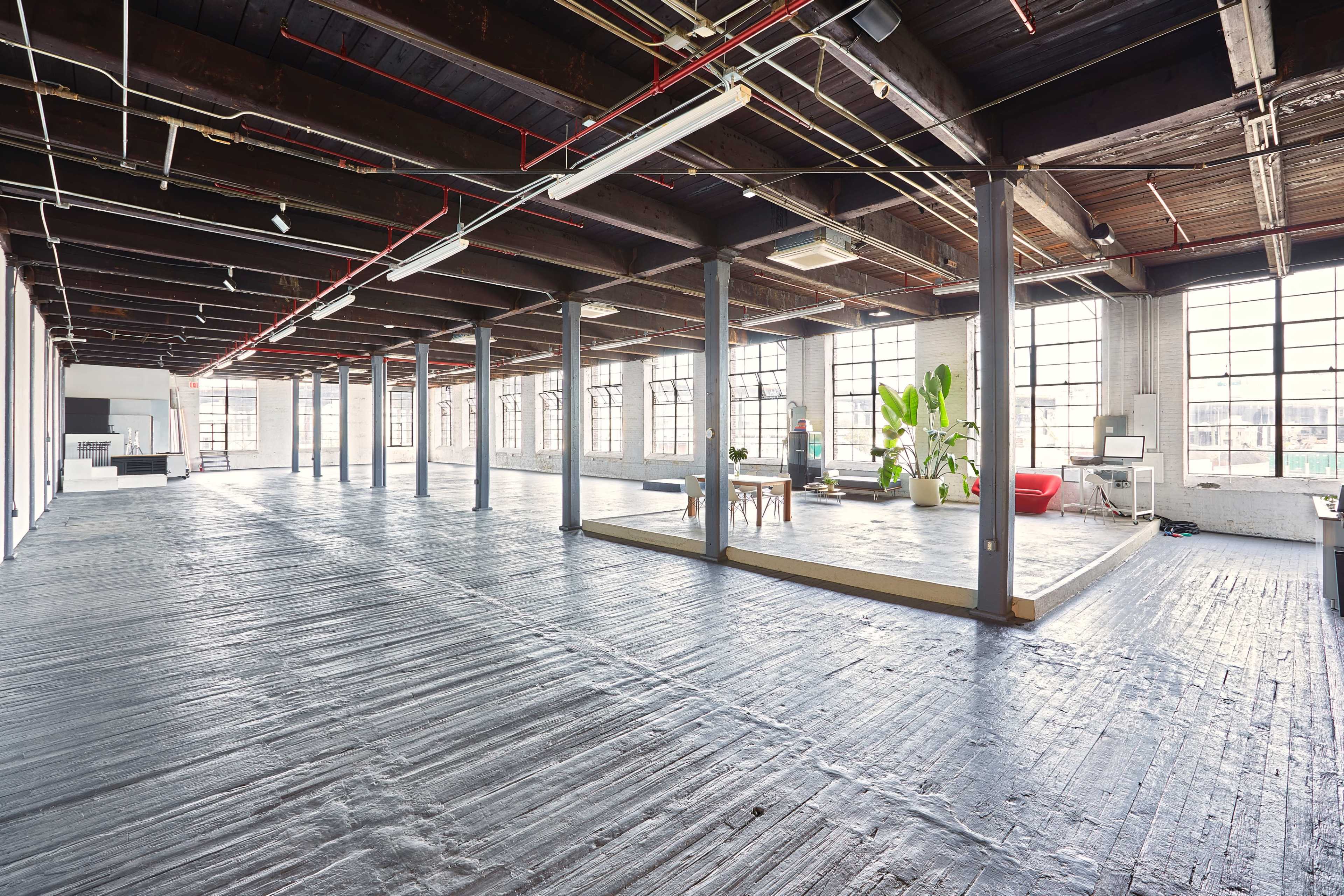A spacious, open-concept loft features large windows, wooden beams, and polished concrete flooring, with distinct areas for lounging and working.