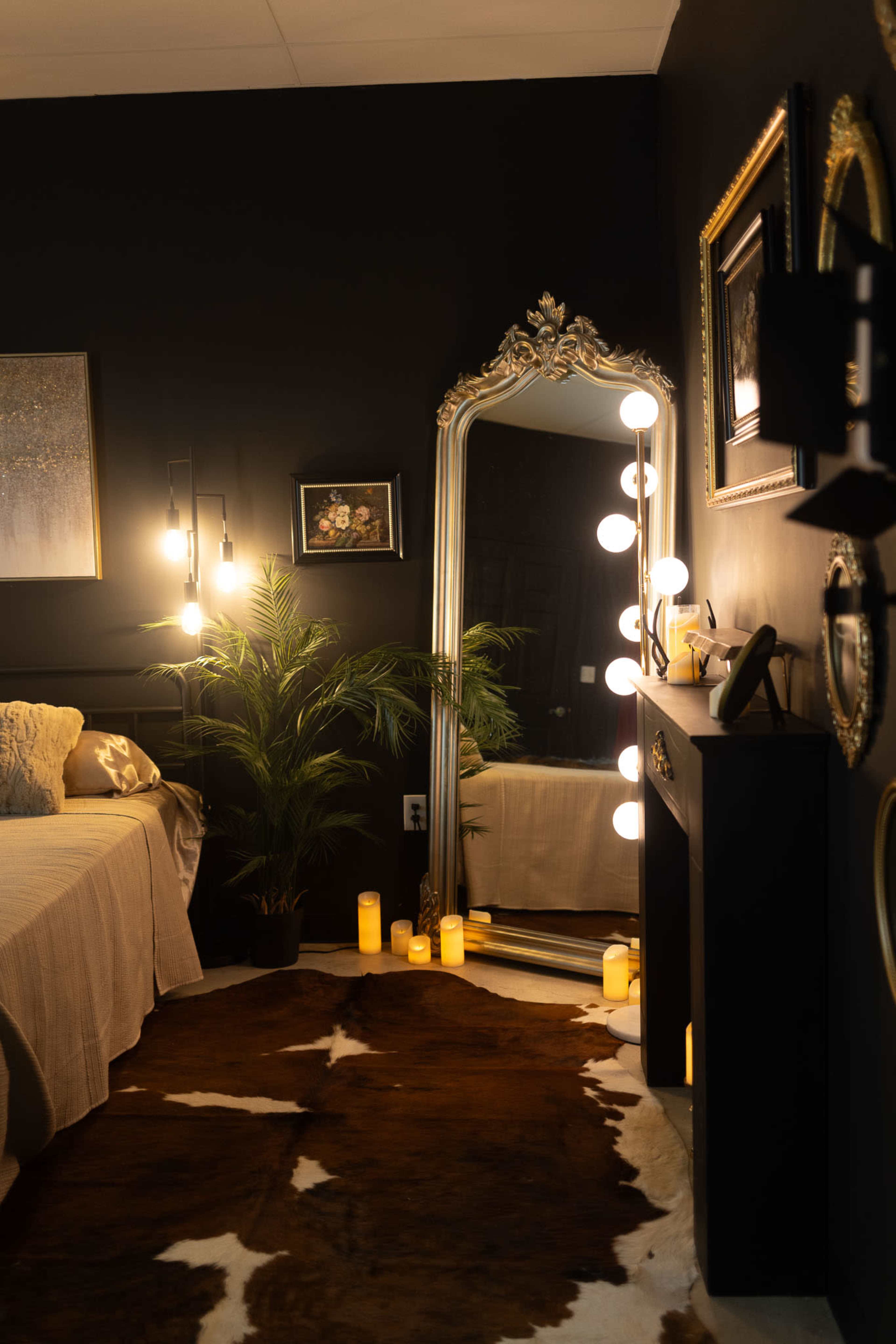 The room features a dark wall with a large mirror, a bed dressed in beige linens, a potted plant, and decorative candles on the floor.