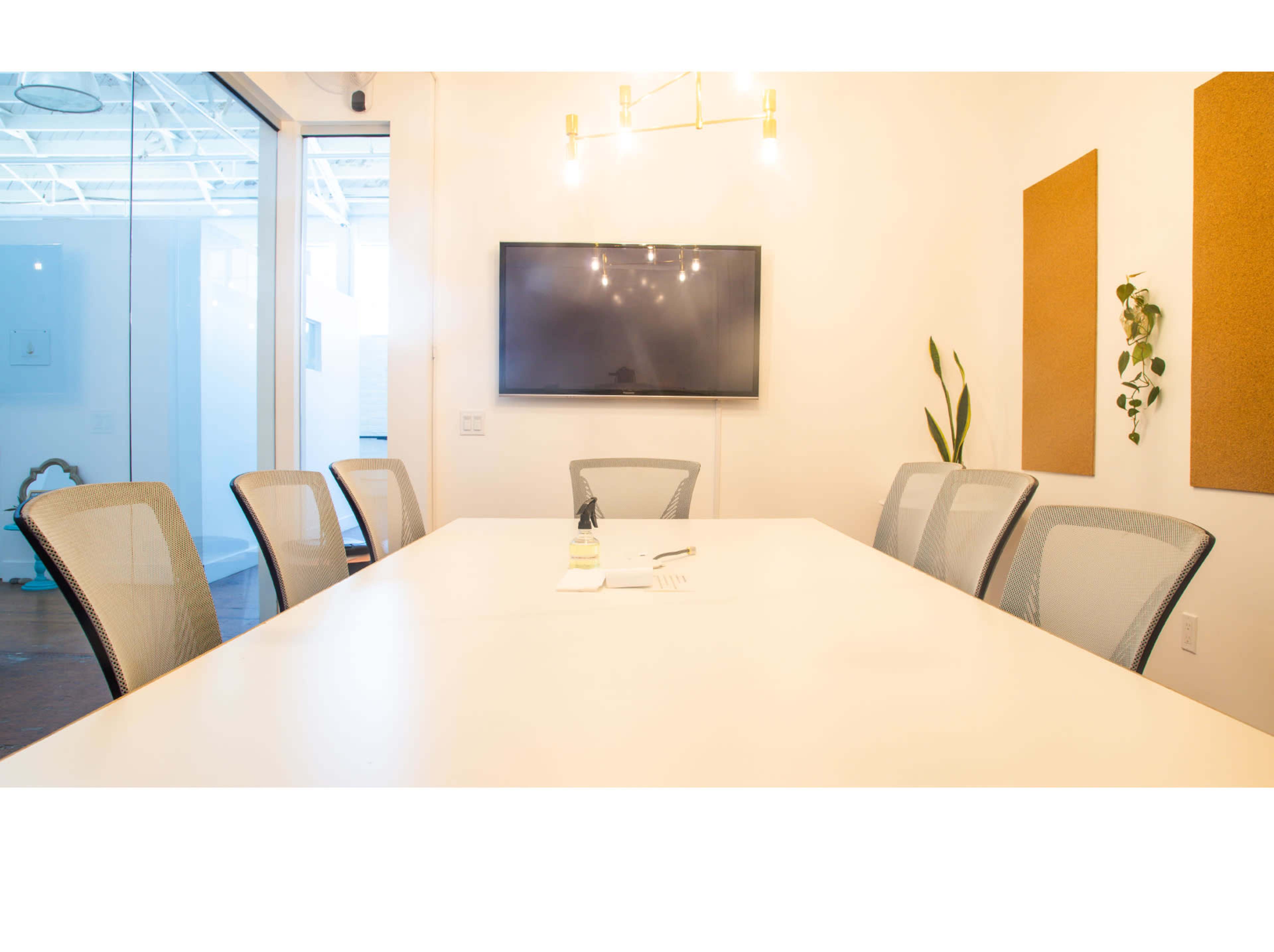 A modern conference room features a long white table surrounded by six mesh chairs, a wall-mounted television, and two cork boards.