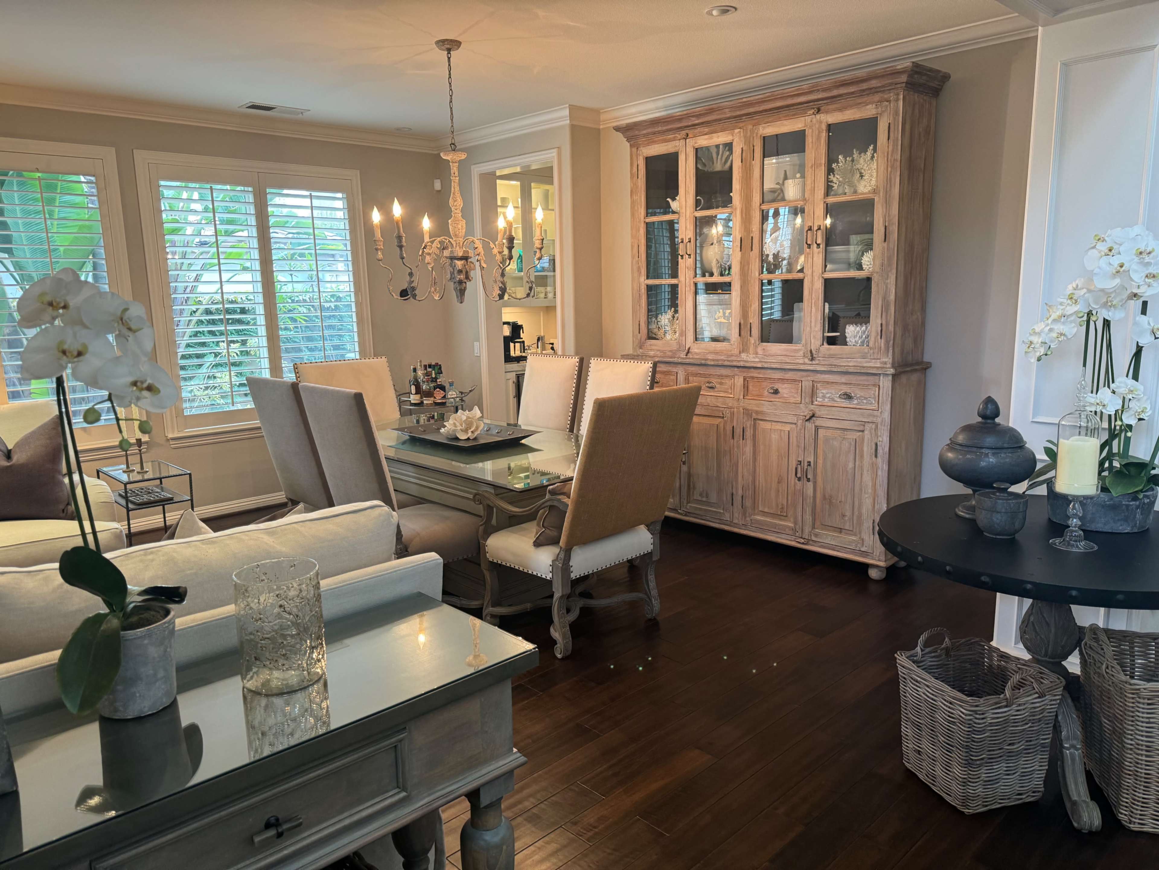 A dining area features a wooden table surrounded by beige chairs, a china cabinet displaying items, and a chandelier hanging from the ceiling, with plants visible through the windows.