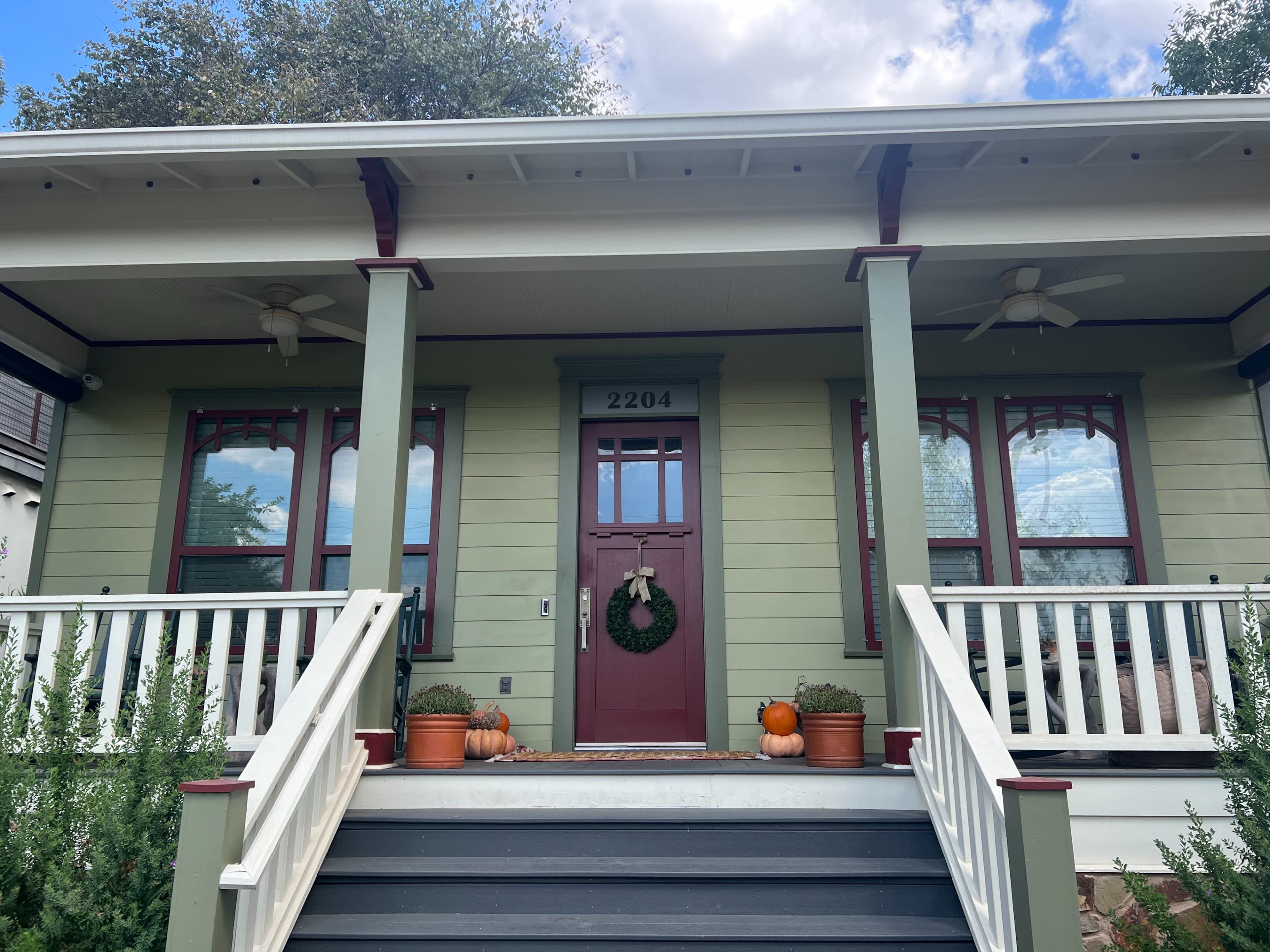 The image shows a green house with a red front door, two fans on the porch ceiling, and decorative pumpkins on the steps.