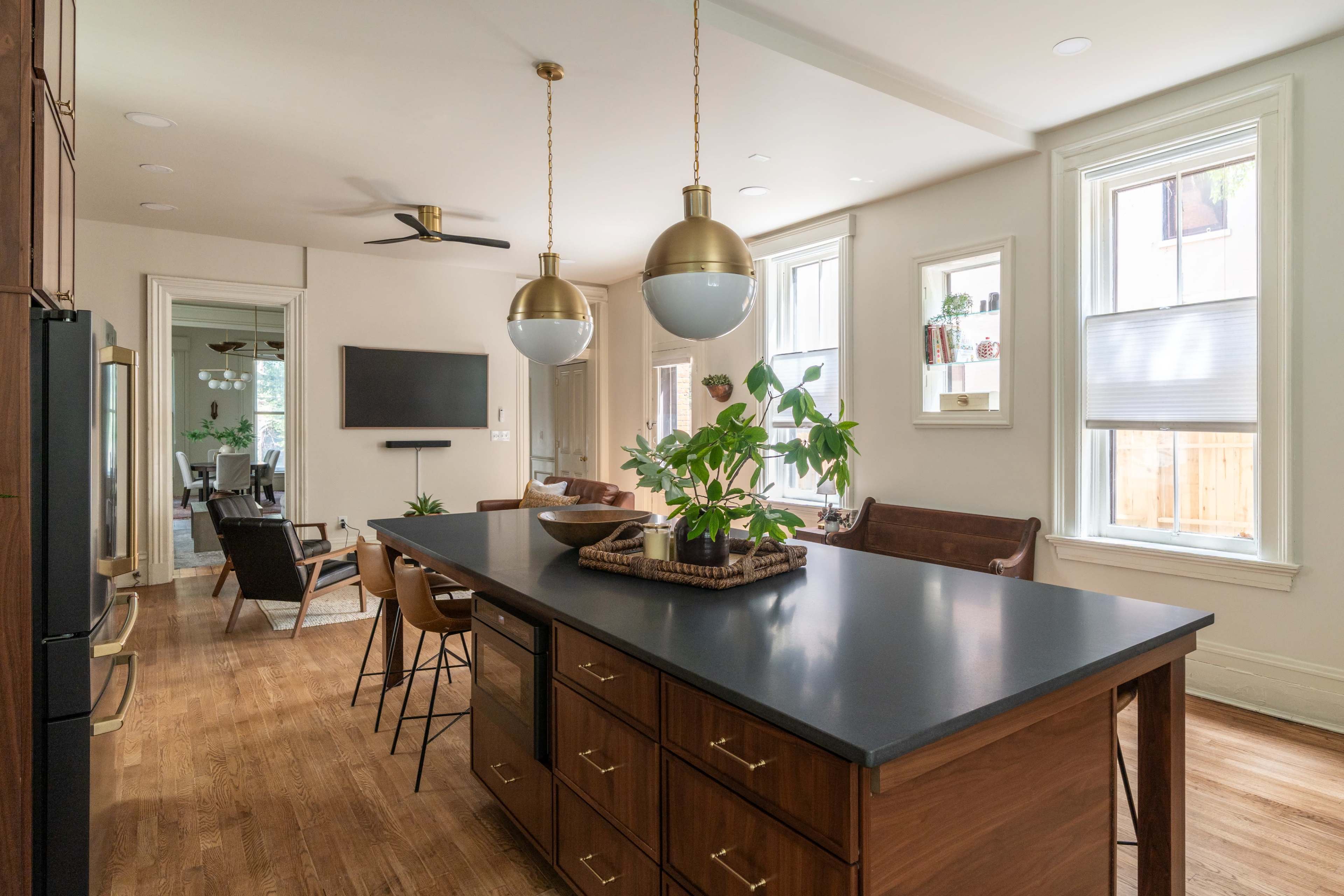 A modern kitchen features a central island with dark cabinetry, gold pendant lights, and a view into a living area with seating.