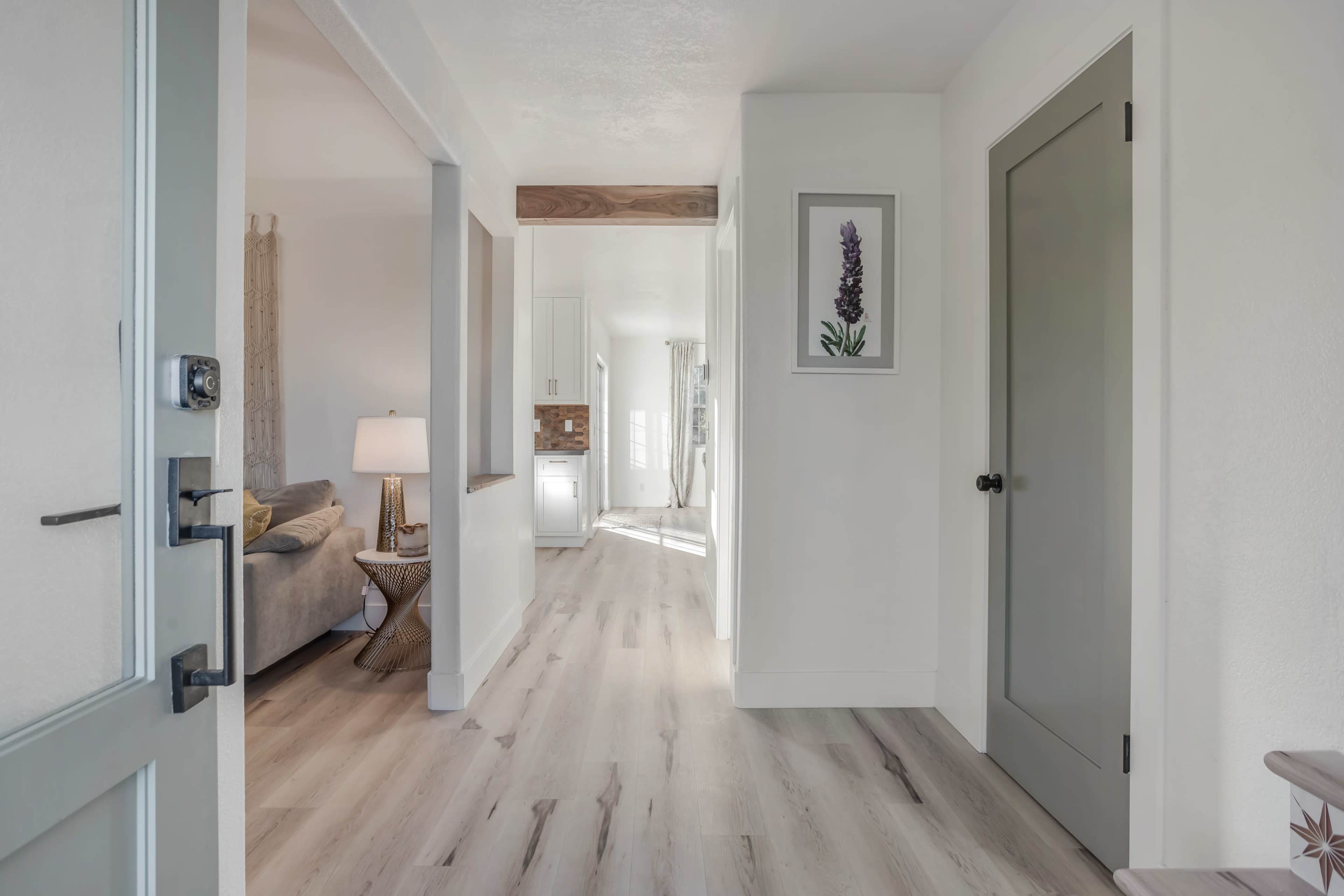The image shows a hallway featuring light wood flooring, a green door, and a view of a living area with a lamp and a framed flower picture on the wall.