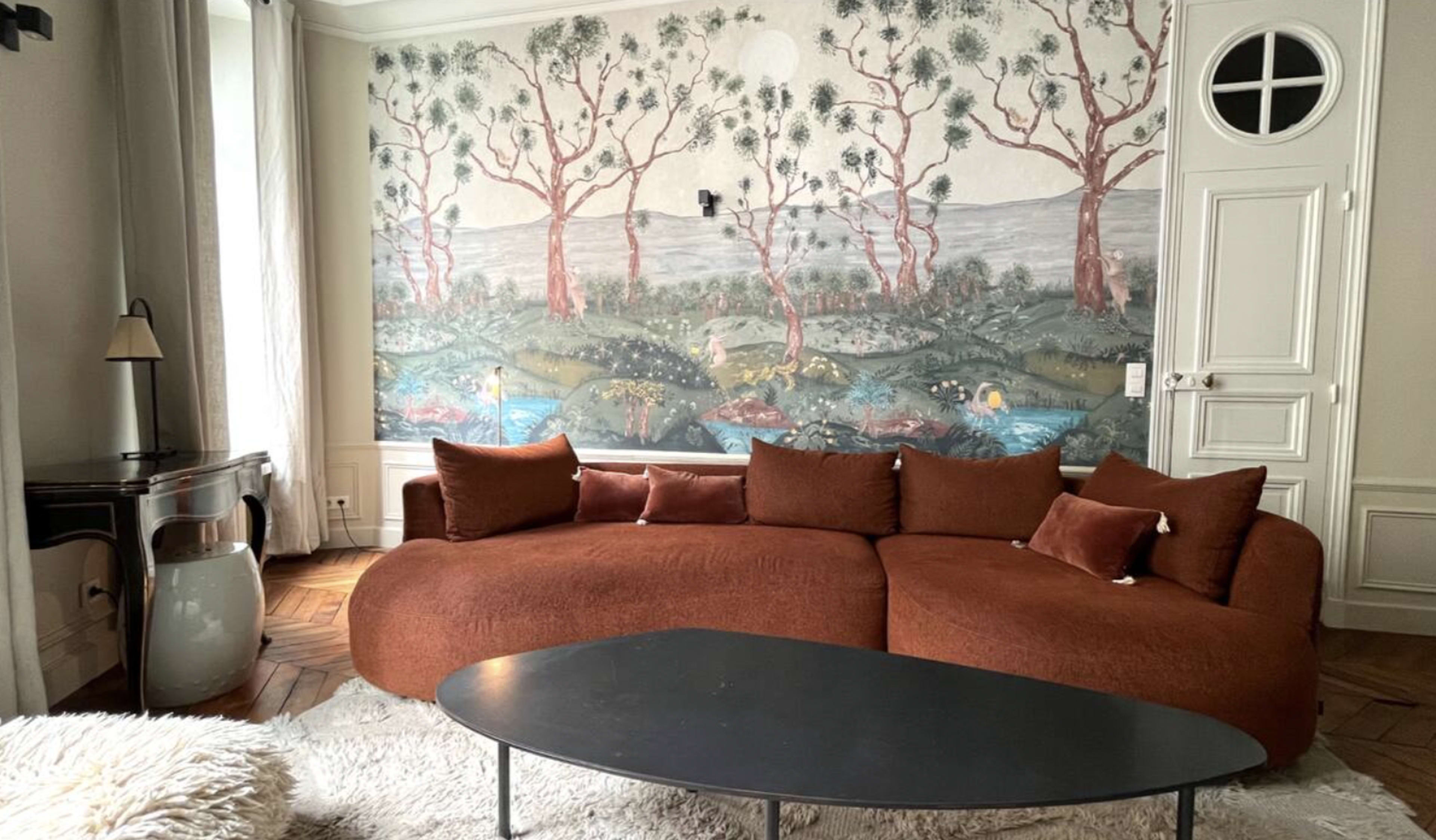 The living room features a curved brown sofa with cushions, a black coffee table, and a vibrant mural of trees and landscape on the wall.