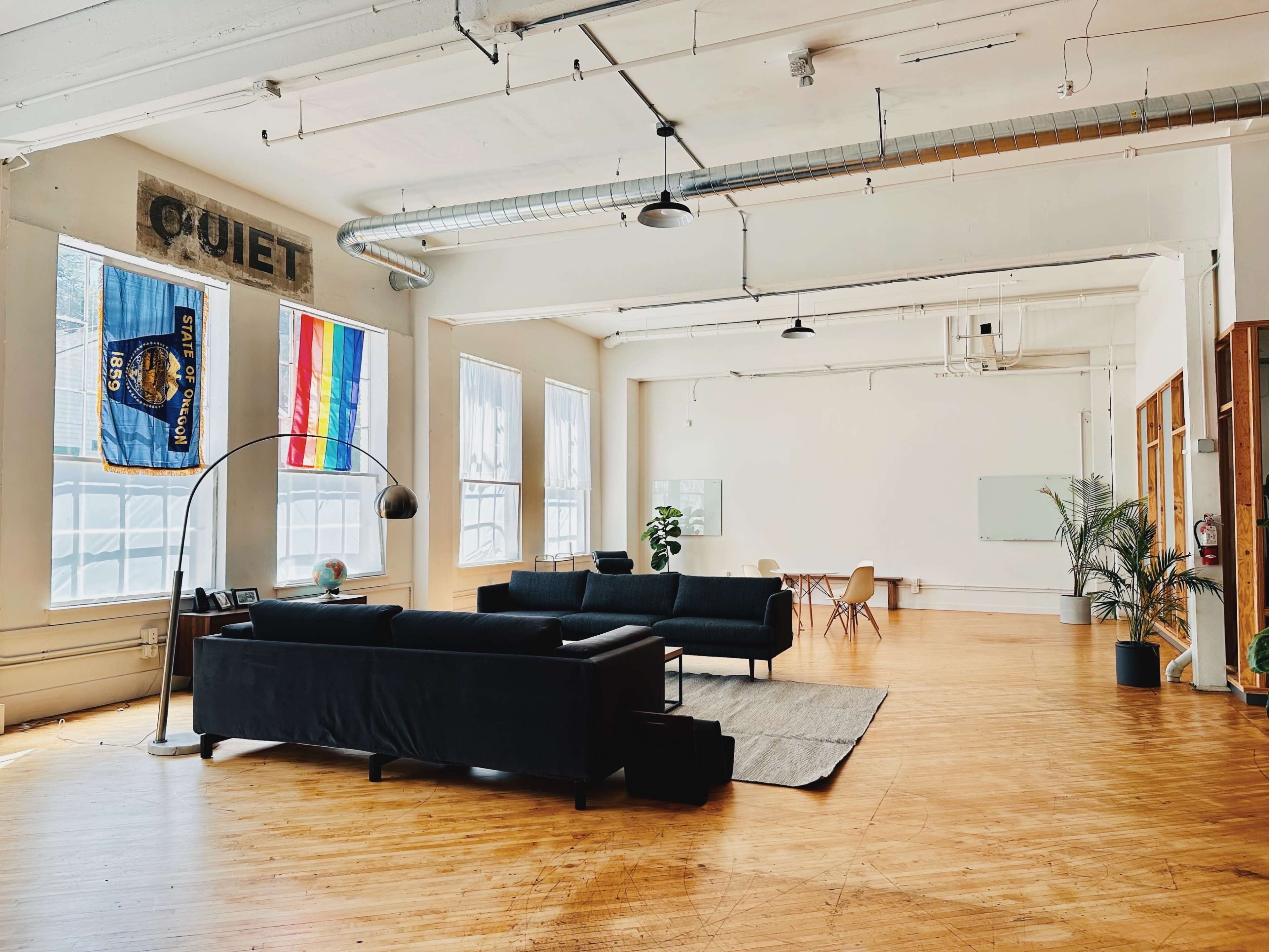 A spacious, minimalist room with two black couches, a floor lamp, a plant, and a meeting area with chairs and a whiteboard.