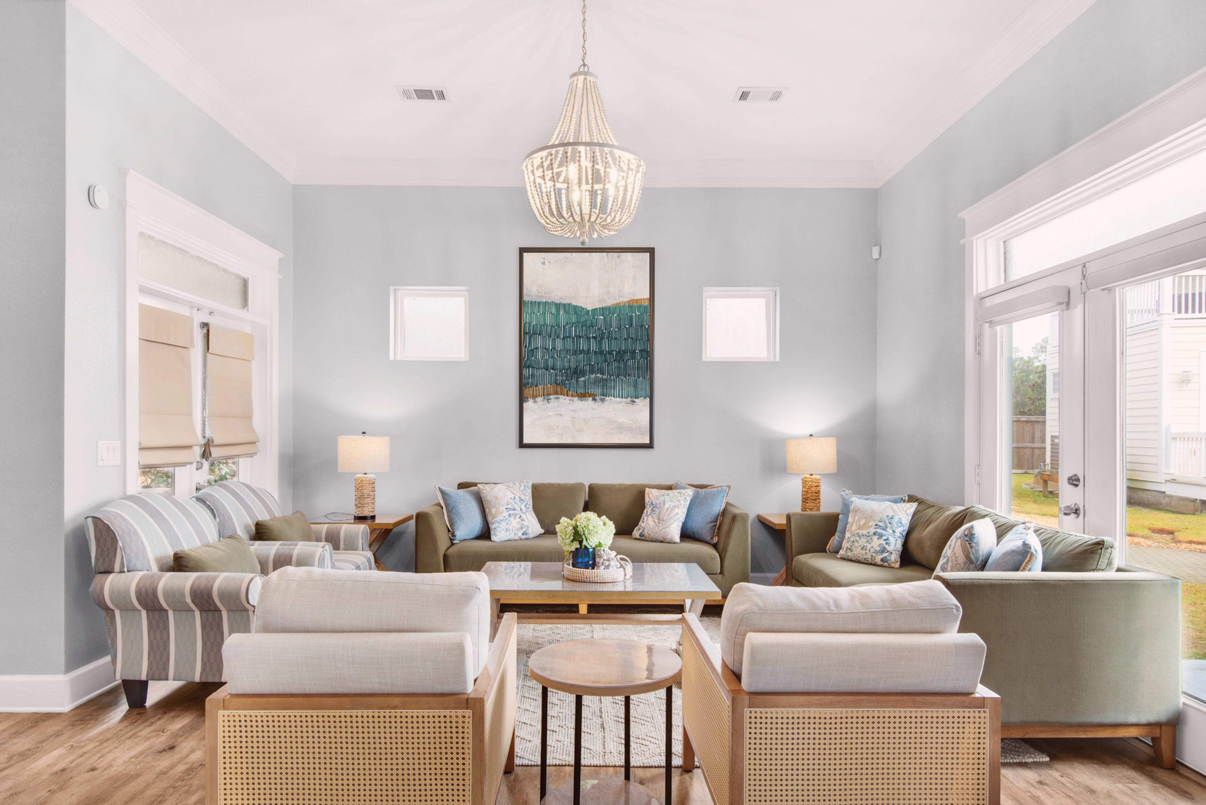 A modern living room features a combination of green and beige seating, a central wooden table, and large windows allowing natural light, adorned with decorative lamps and an art piece on the wall.
