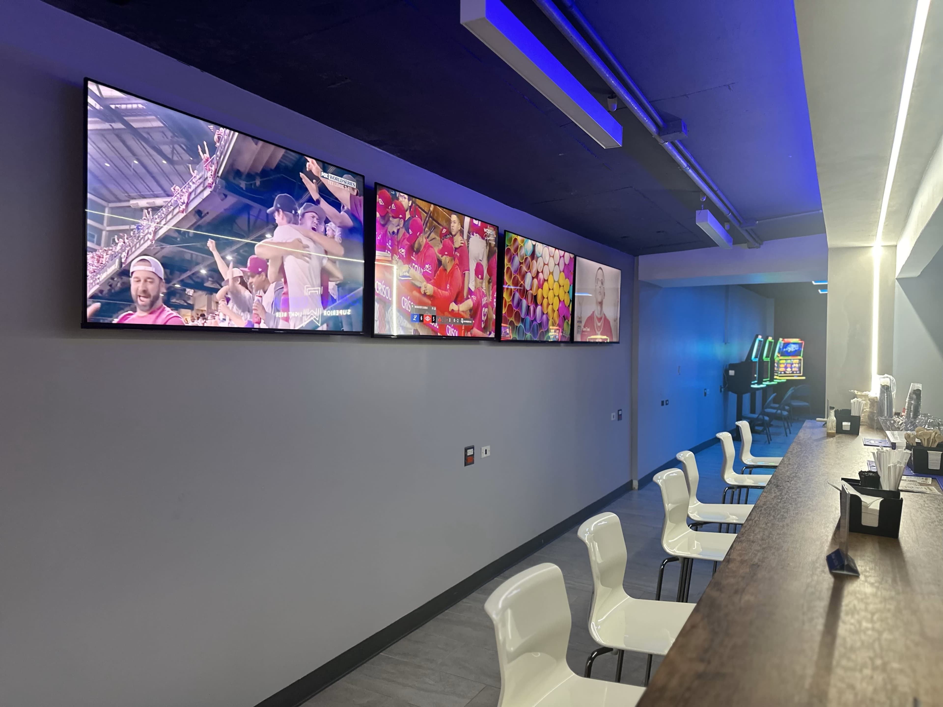 The image shows a modern bar area with several large screens displaying colorful video content, accompanied by a row of white chairs and a wooden counter.