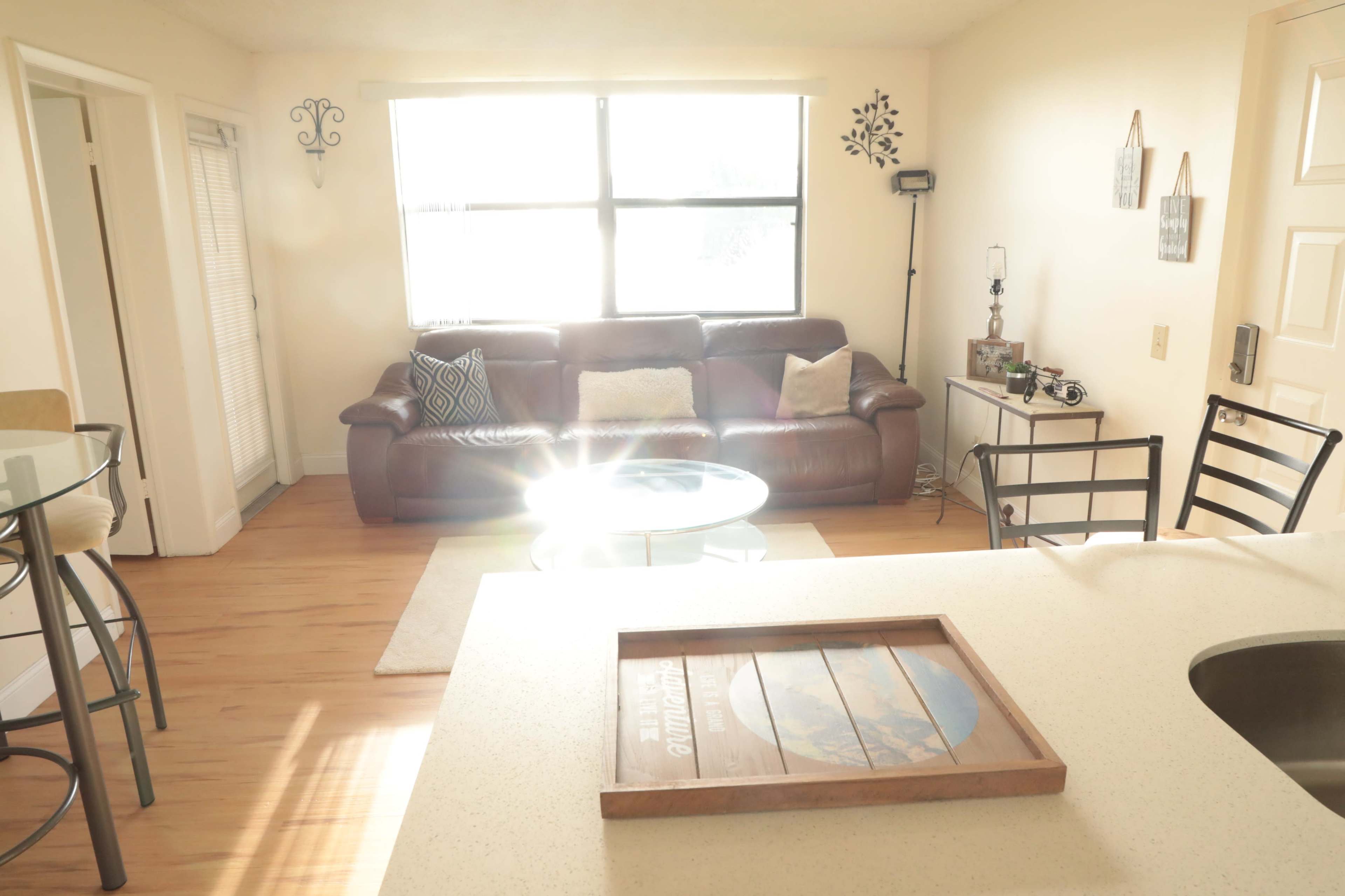 A bright living room with a brown leather couch, a glass-top dining table, and light wood flooring.