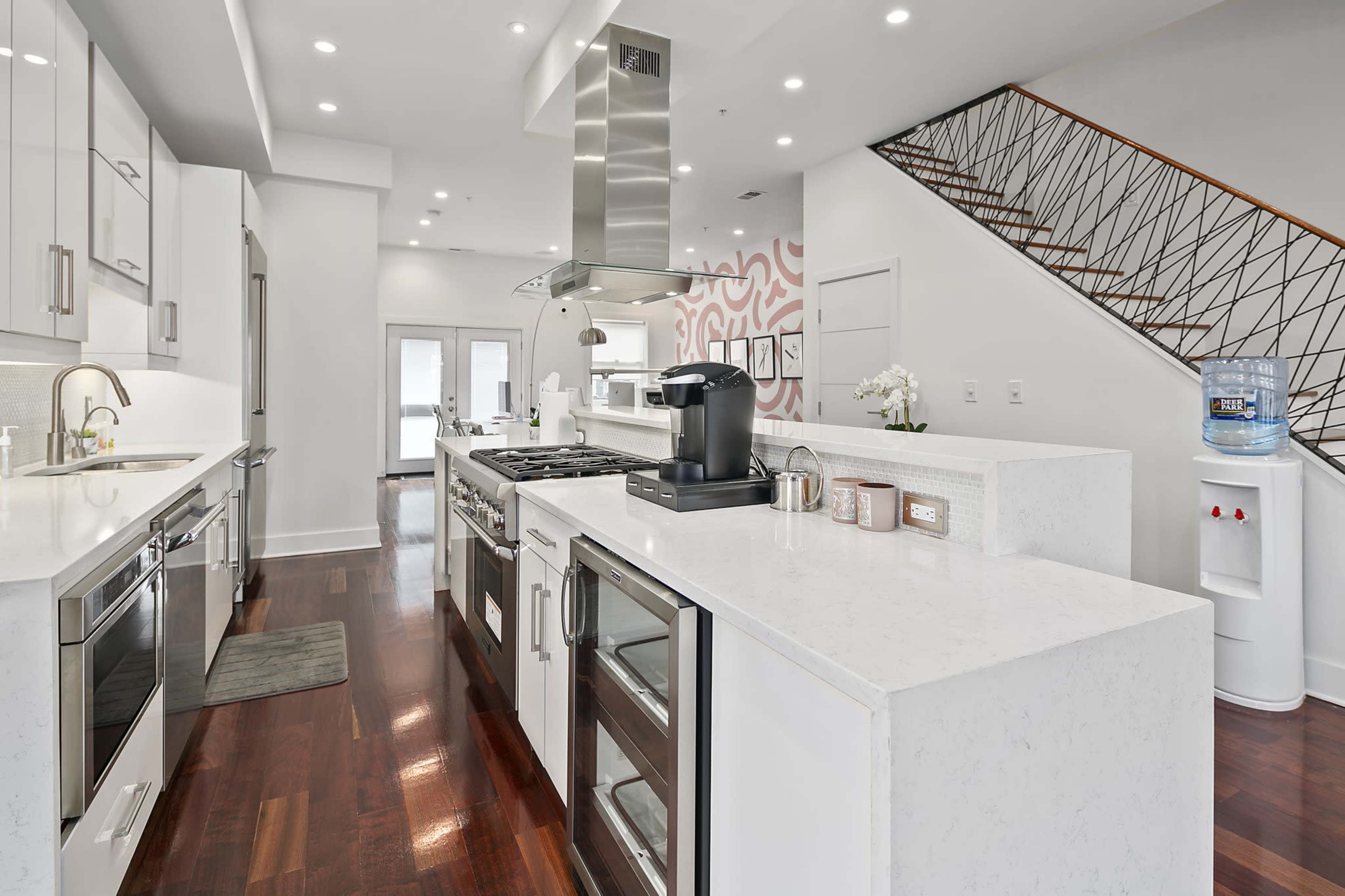 A modern kitchen featuring a large island with a cooktop, stainless steel appliances, and a staircase in the background.