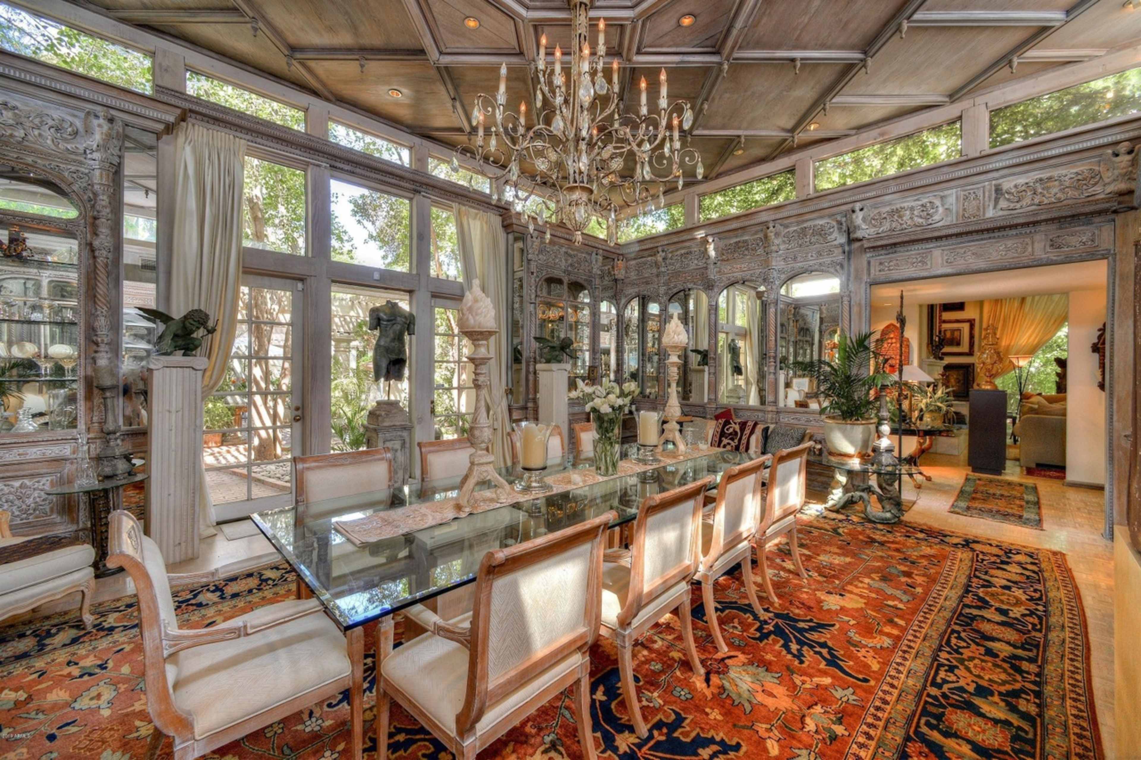 The image shows an elegant dining room featuring a large glass table surrounded by upholstered chairs, ornate wooden detailing on the walls, a chandelier, and large windows providing natural light.