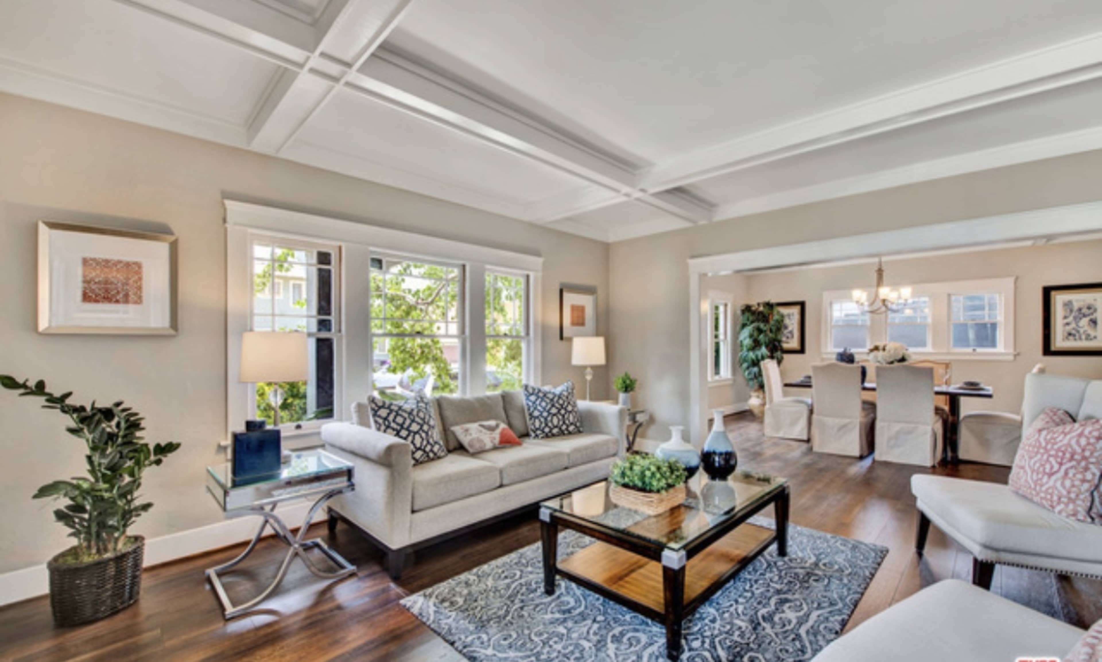 A modern living room features a light-colored sofa, a coffee table, and large windows allowing natural light to flow into the space, with a dining area visible in the background.