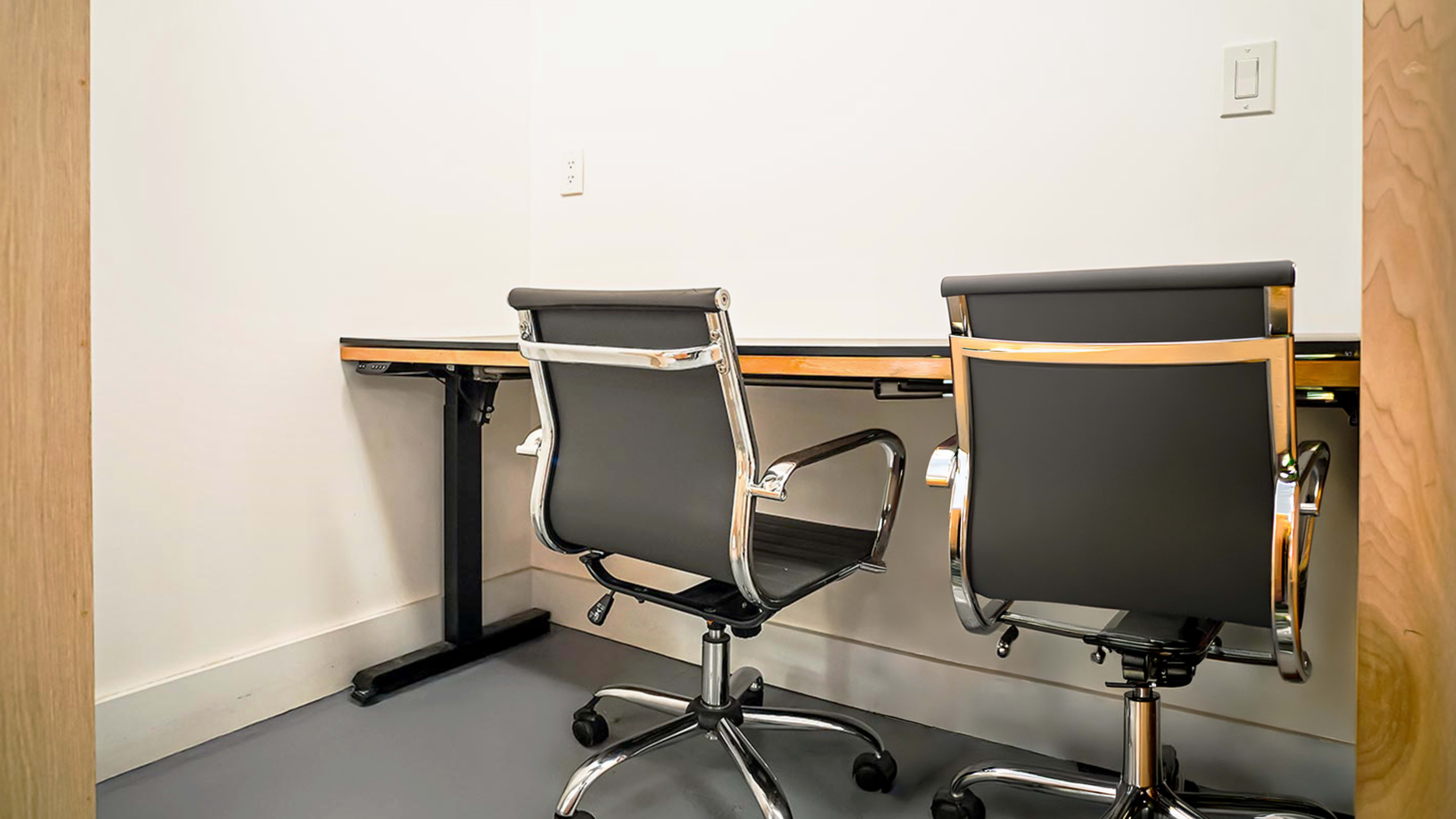The image shows a small office space with two black office chairs positioned at a wooden desk against a plain white wall.