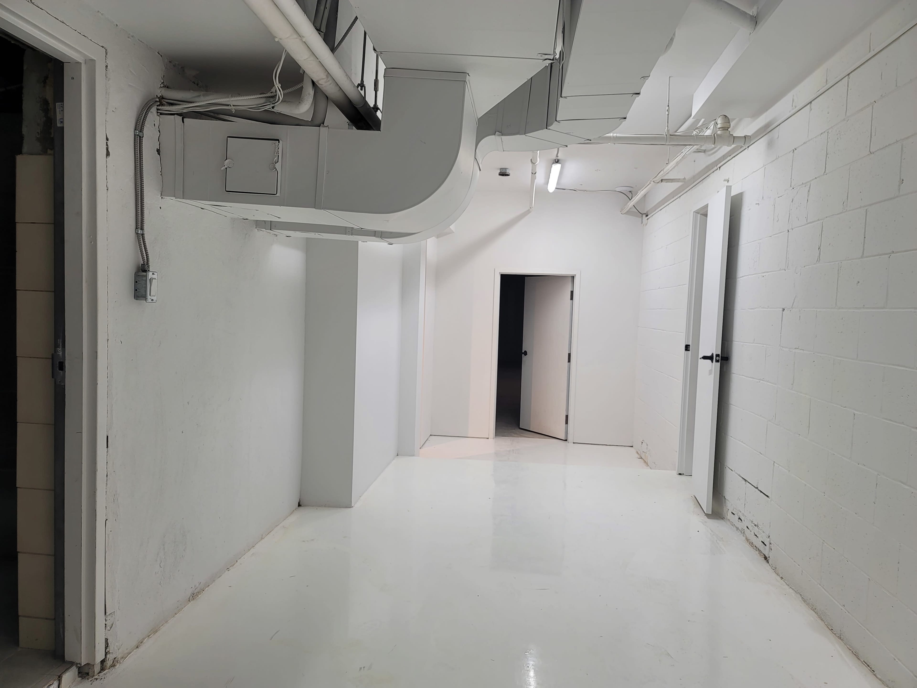 A light-colored corridor in a basement with several doors and exposed ductwork on the ceiling.