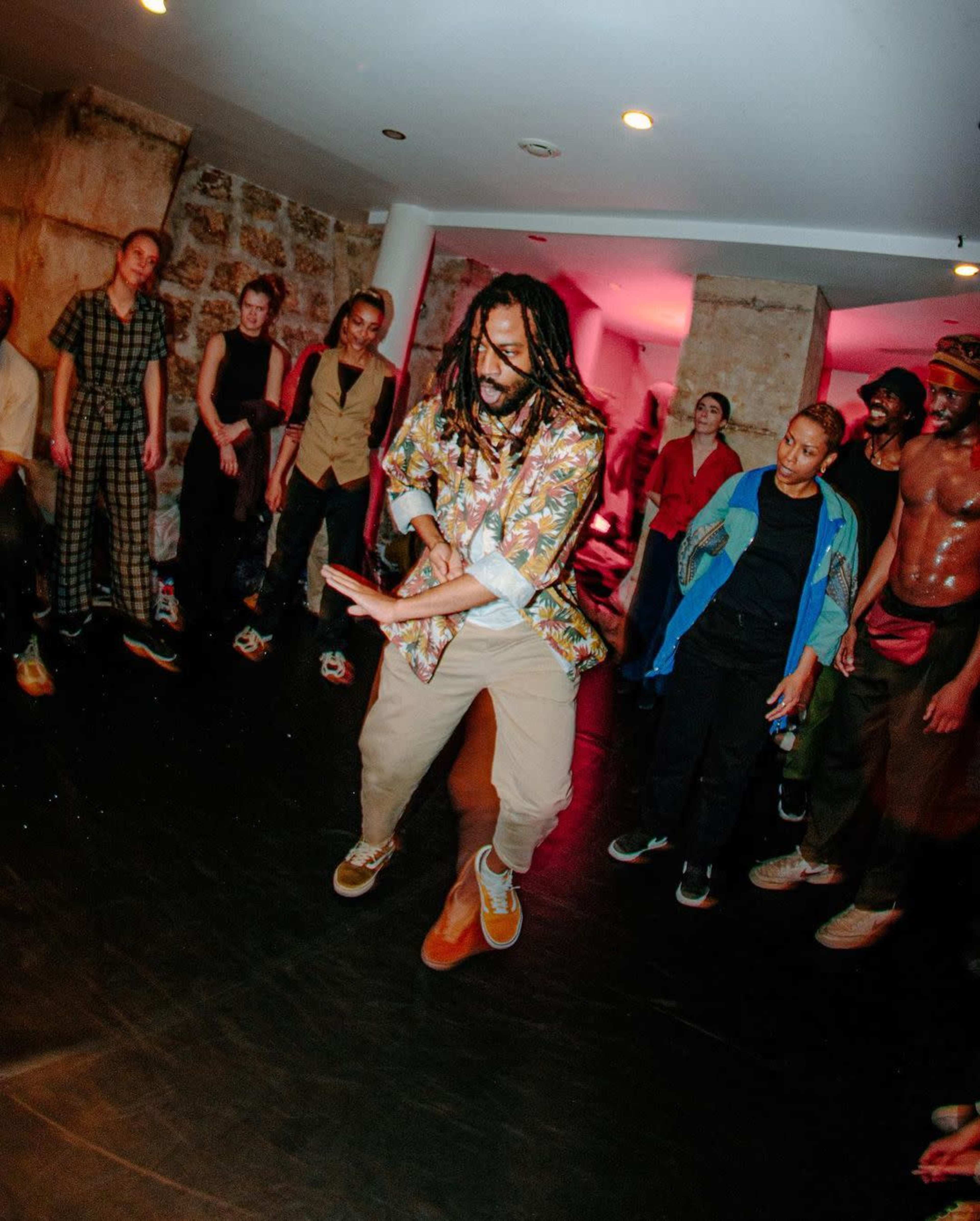 A group of people observes a dancer performing in a dimly lit room with a textured stone wall.