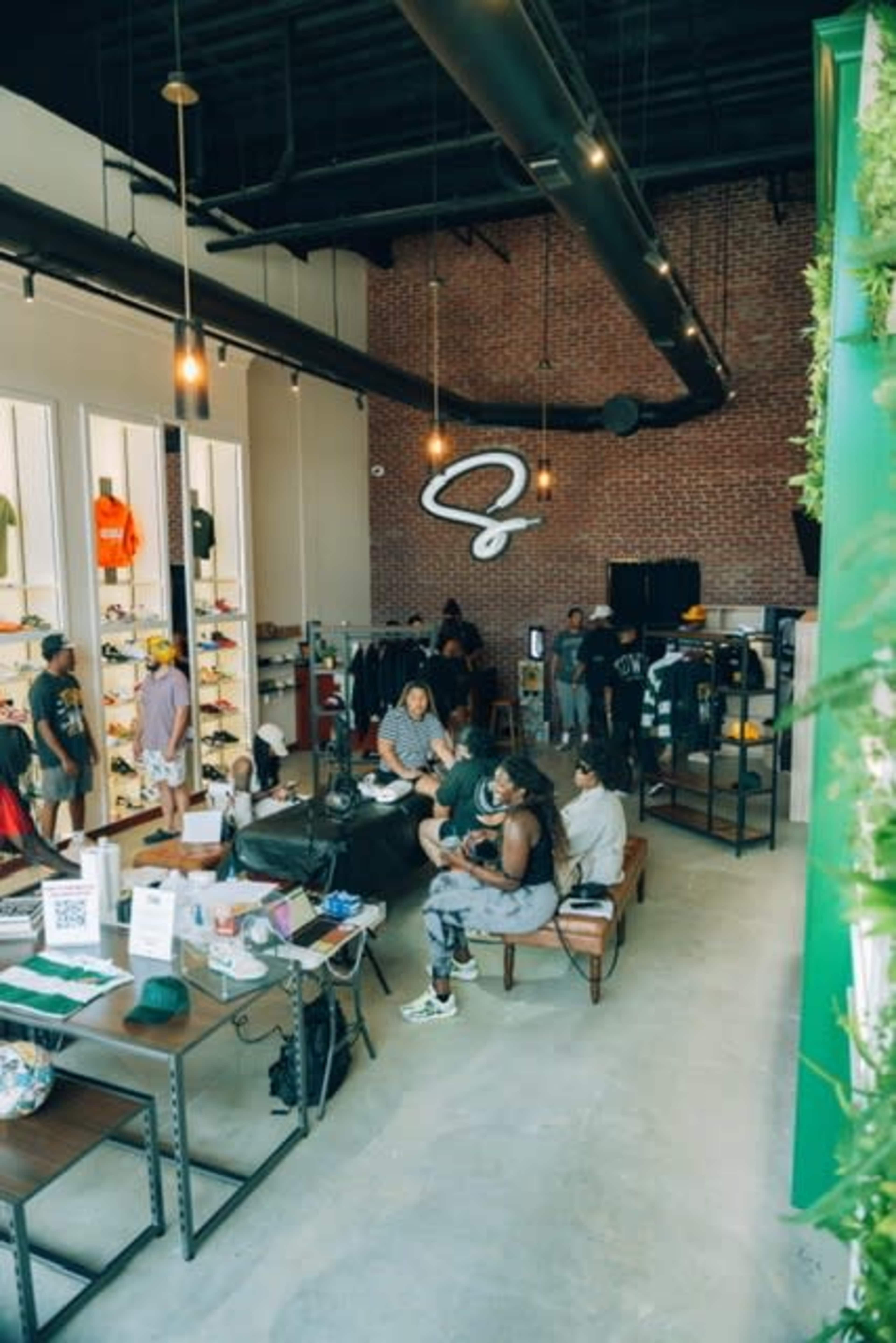A group of people is gathered inside a trendy retail store featuring clothing and shoes displayed on shelves against a brick wall.