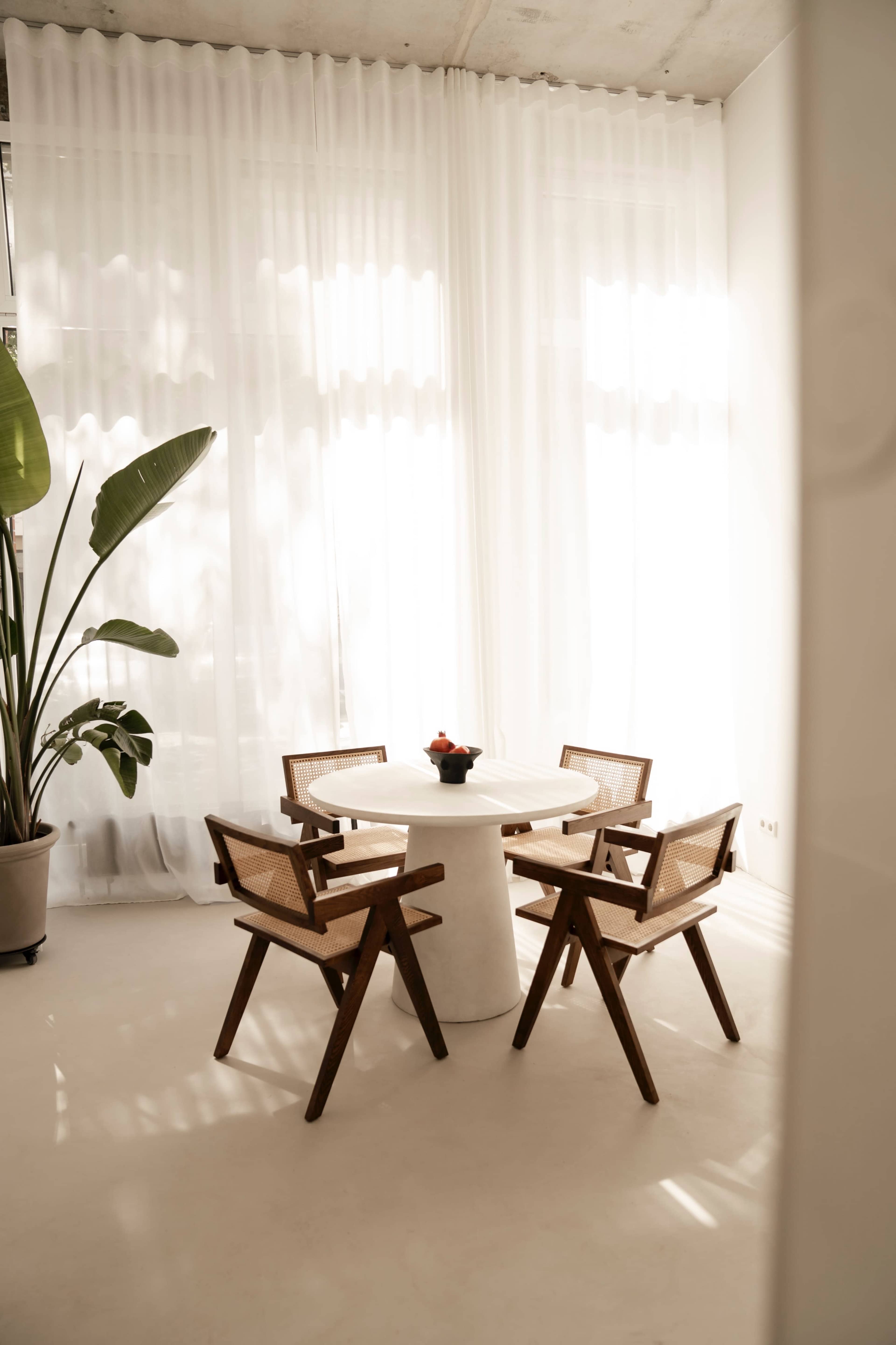 A round white table surrounded by four wooden chairs is positioned near a large window with sheer curtains and a potted plant.