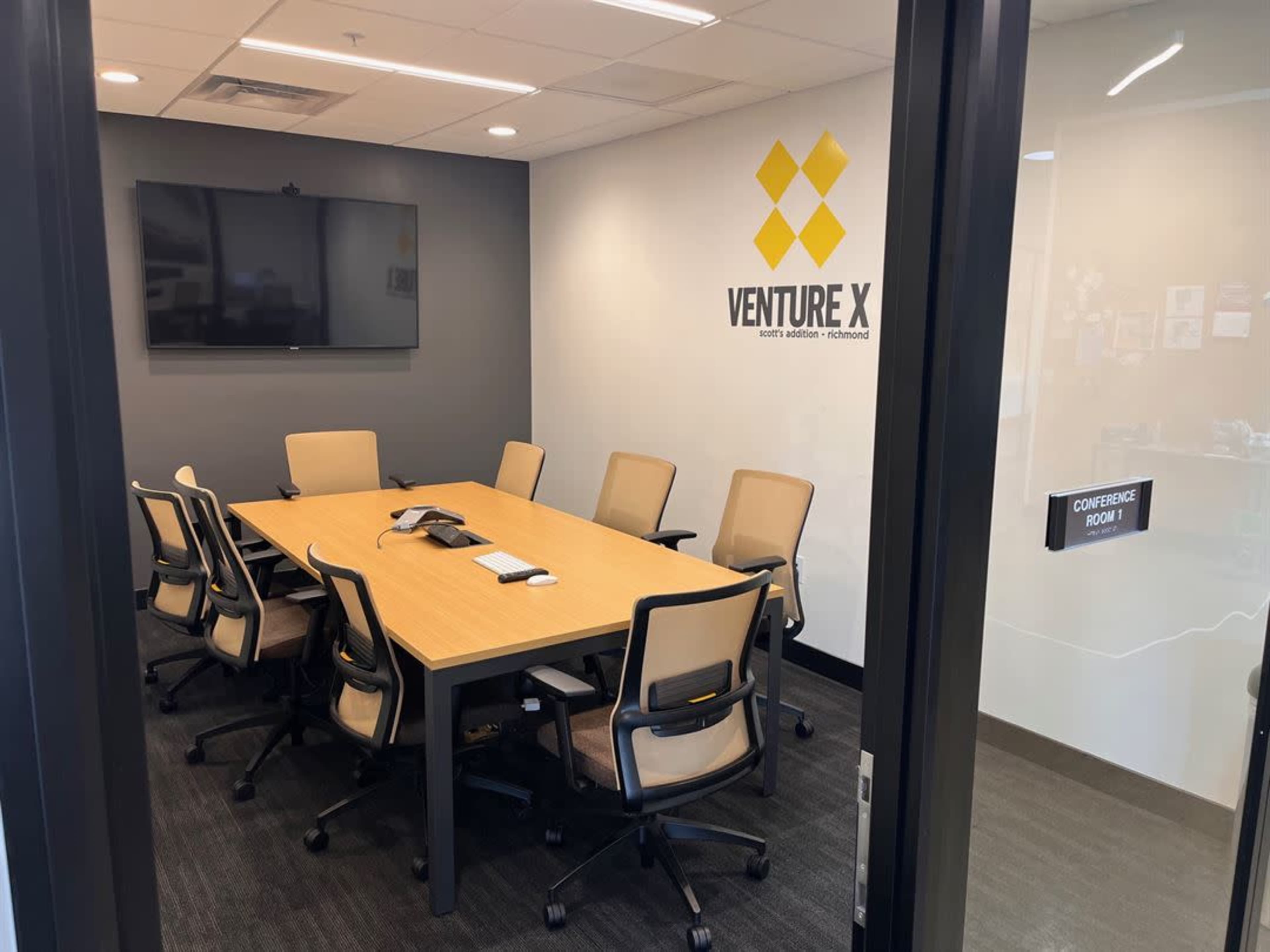 A conference room equipped with a long table, eight chairs, a large wall-mounted screen, and a phone on the table.