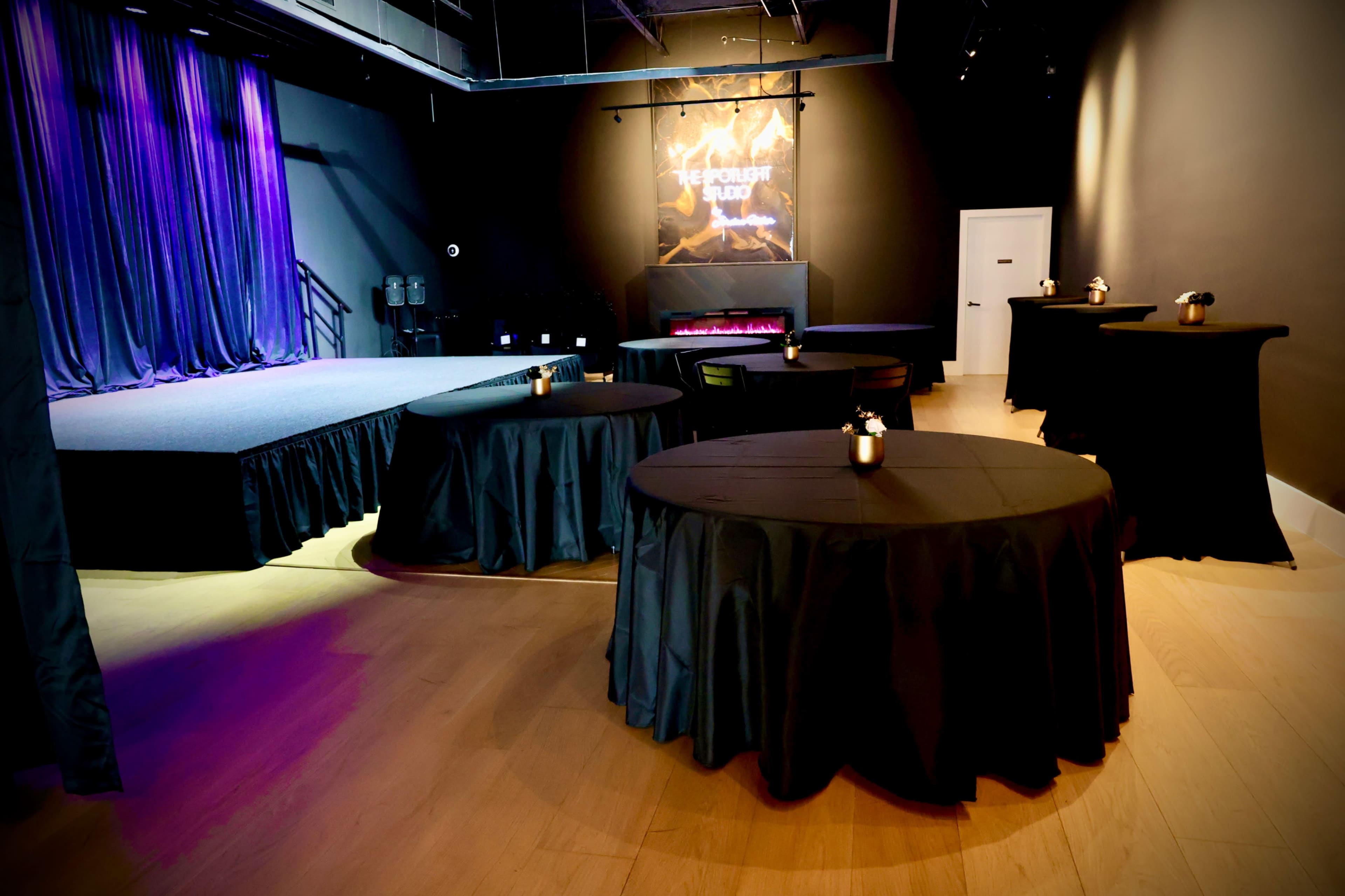 The image shows an indoor event space featuring a stage with purple curtains, a lit backdrop, and several round tables set with black tablecloths and candles.