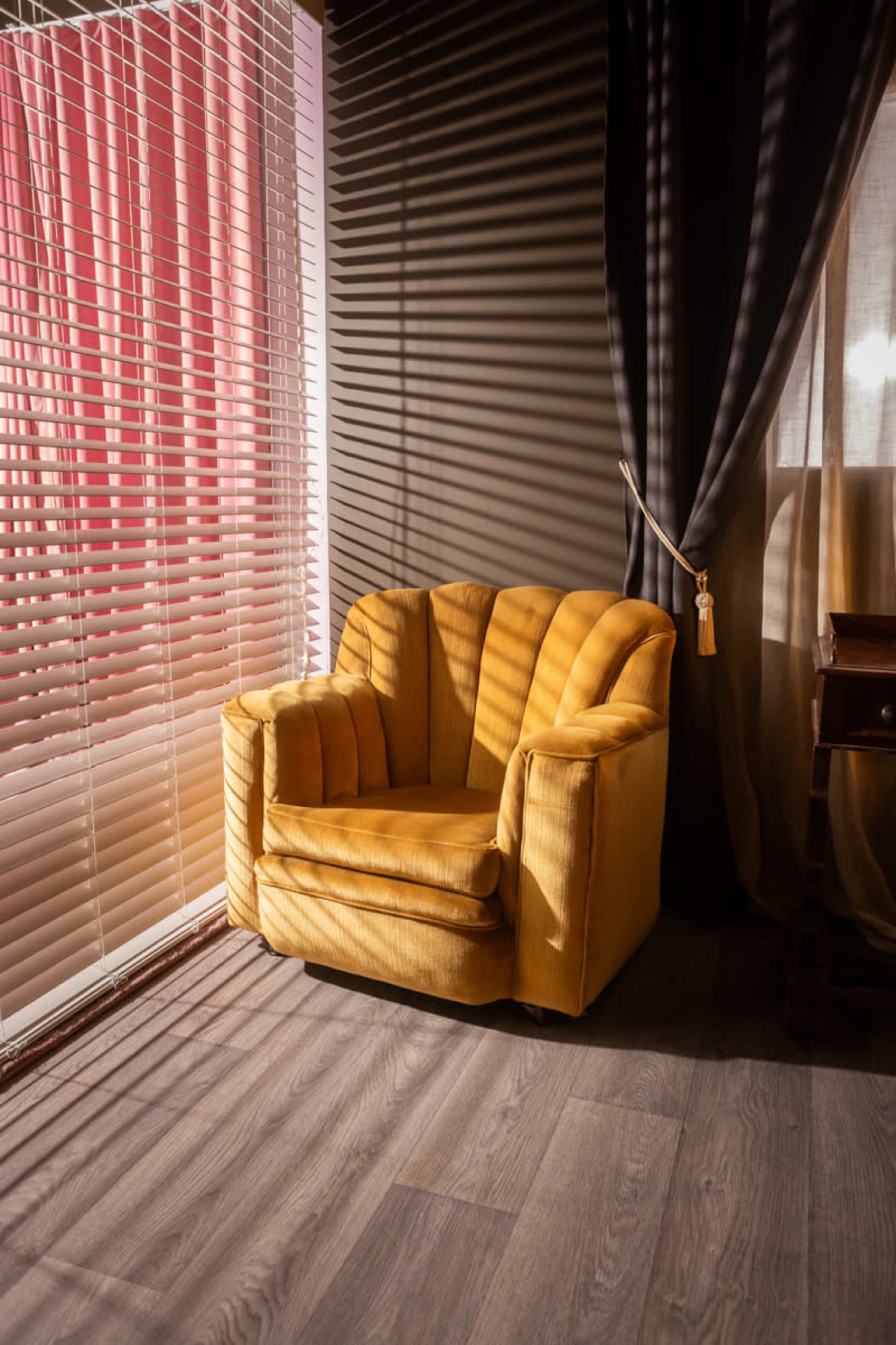 A yellow armchair is positioned beside a window with partially closed blinds, casting striped shadows on the wooden floor.