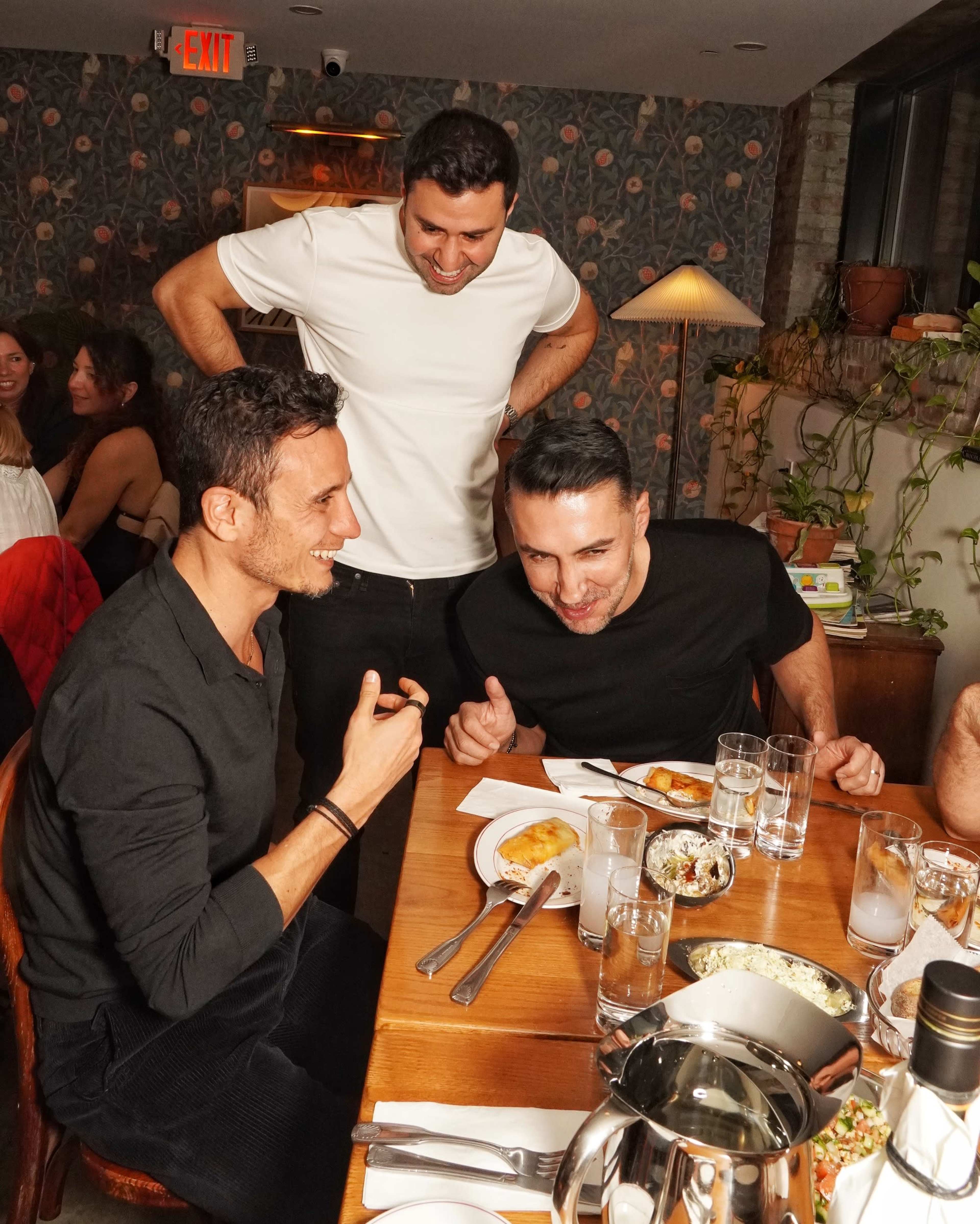 Three men are gathered around a wooden dining table, laughing and engaging in conversation while enjoying their meal in a restaurant setting with a lively atmosphere.