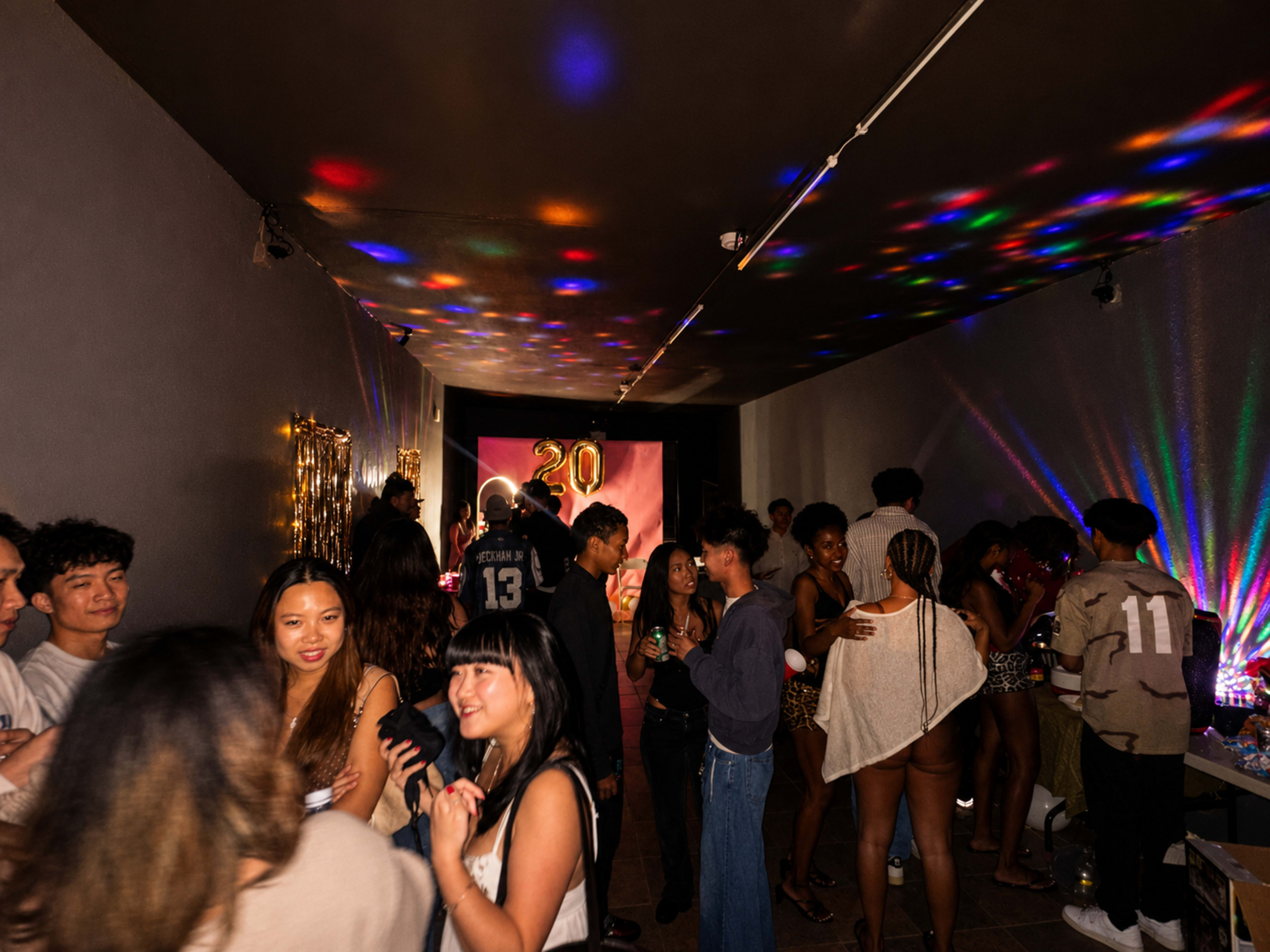 A vibrant party scene is filled with diverse people socializing in a dimly lit room adorned with colorful lights and decorations.