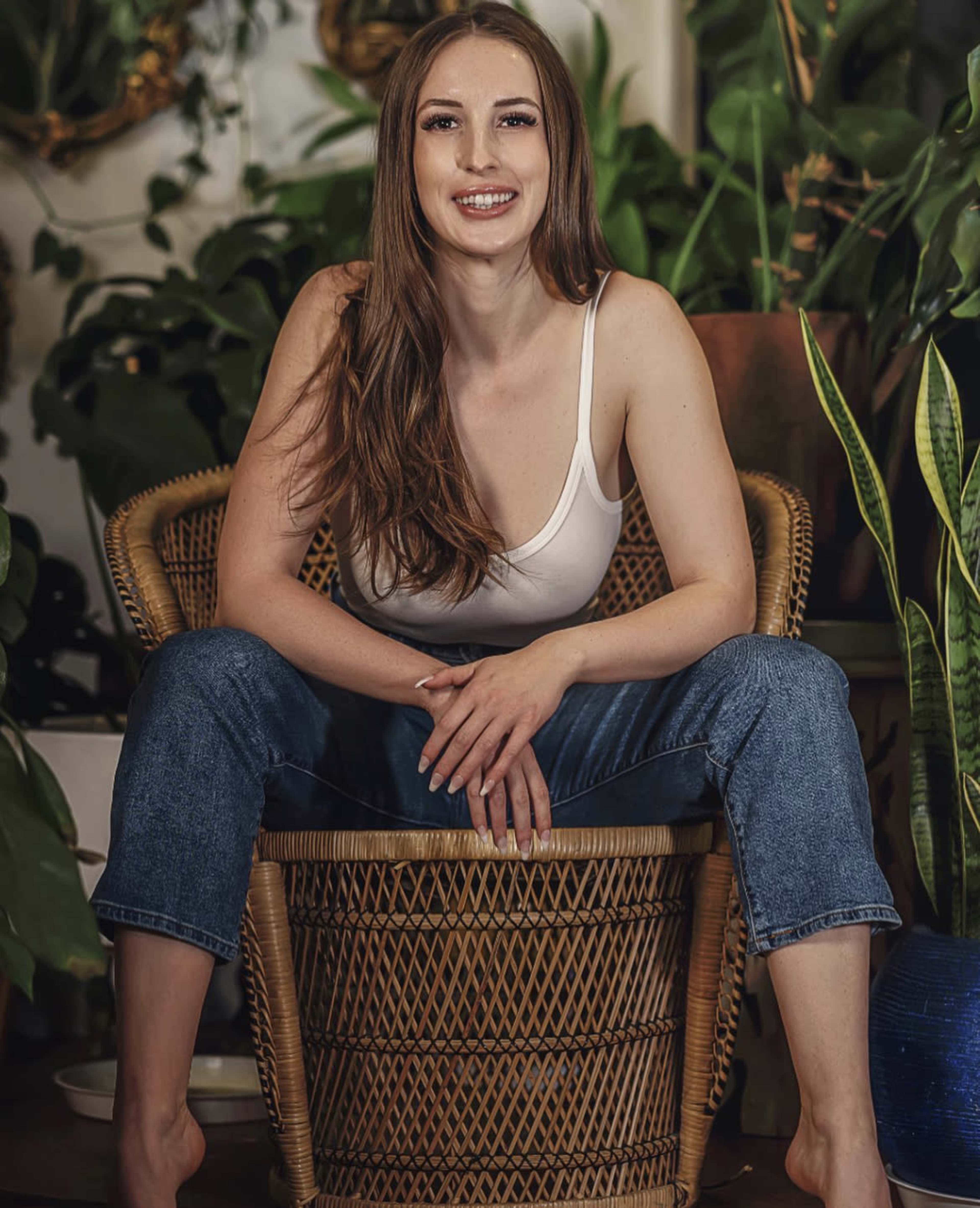 A woman sits cross-legged in a wicker chair surrounded by various indoor plants.
