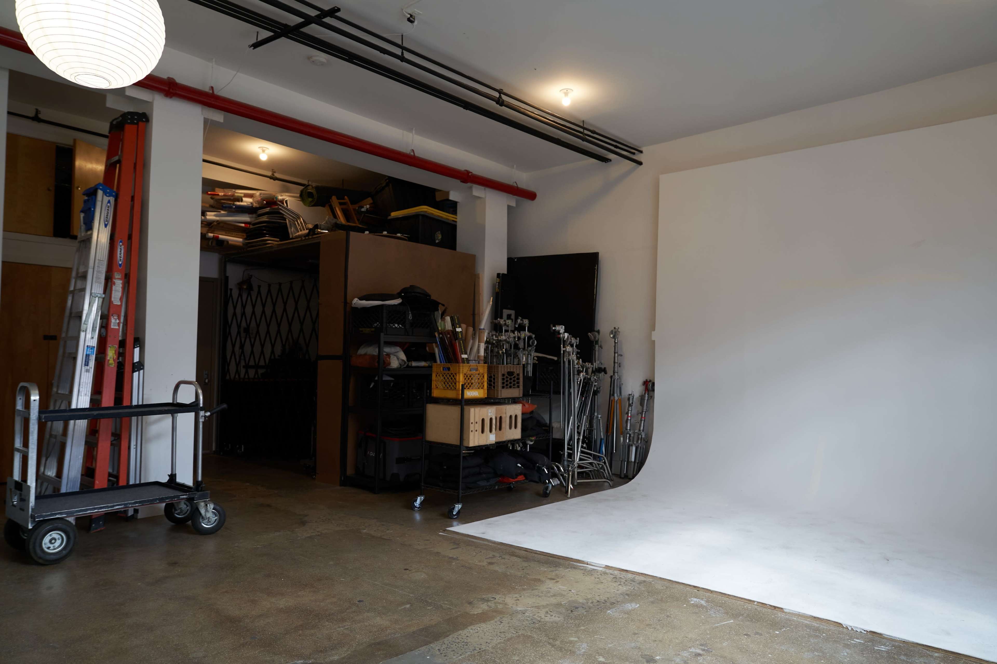 The image shows a spacious studio with a white backdrop, a cart on the left, and storage areas filled with equipment and supplies in the background.