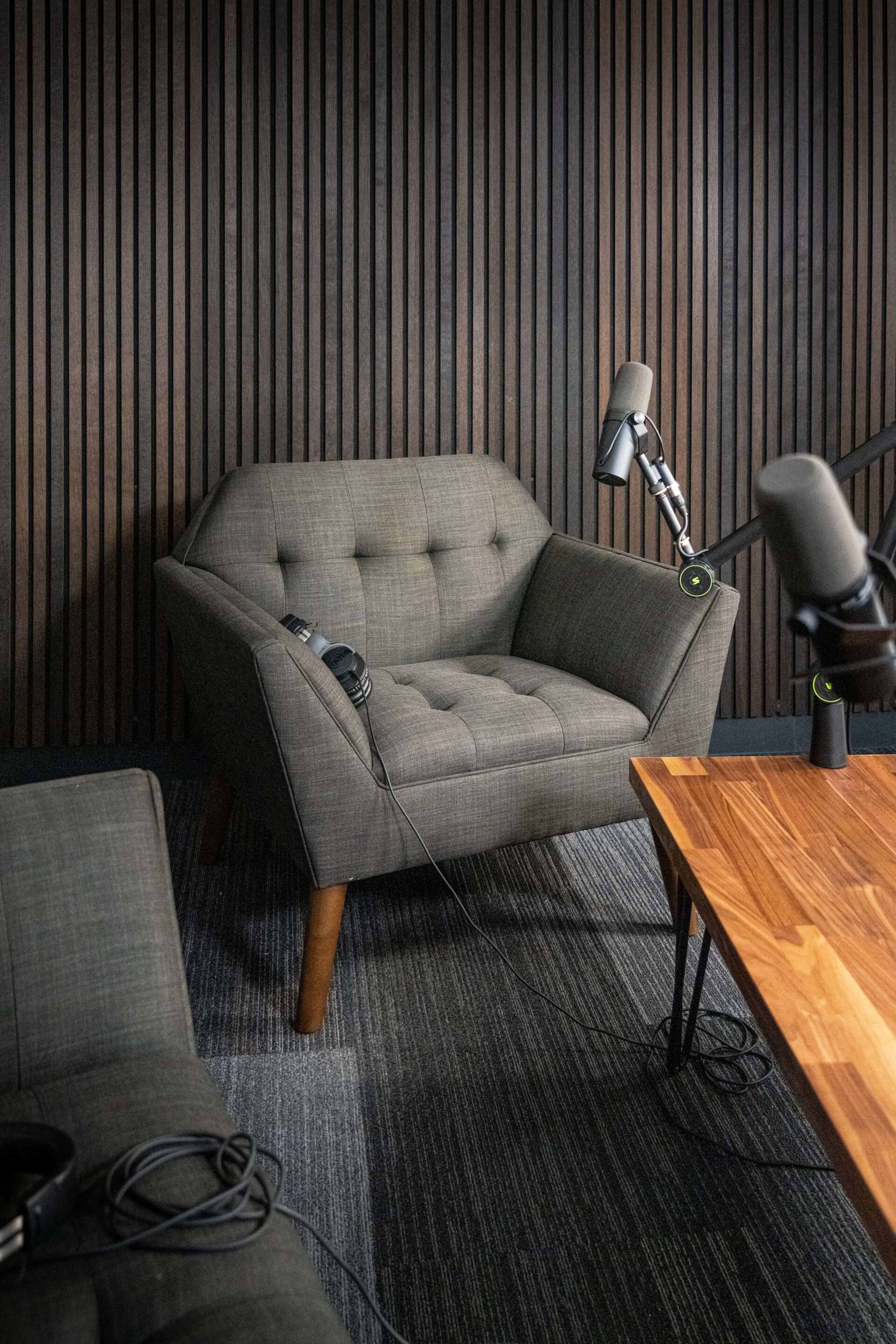 A modern recording studio setup with a green armchair, a wooden table, and microphone stands positioned nearby.