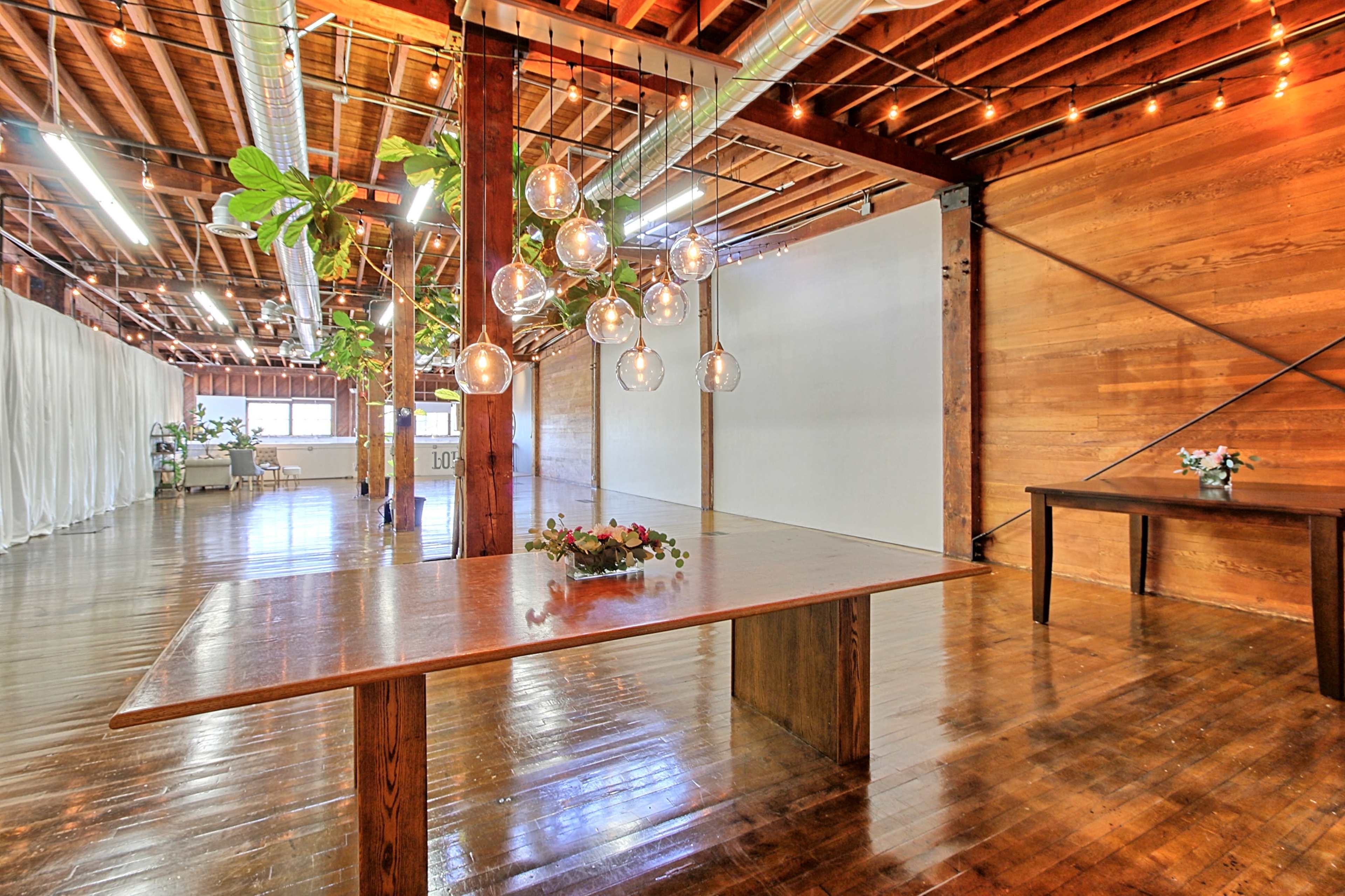 A spacious room with wooden beams, polished floors, and a large table adorned with a floral centerpiece under hanging glass light fixtures.