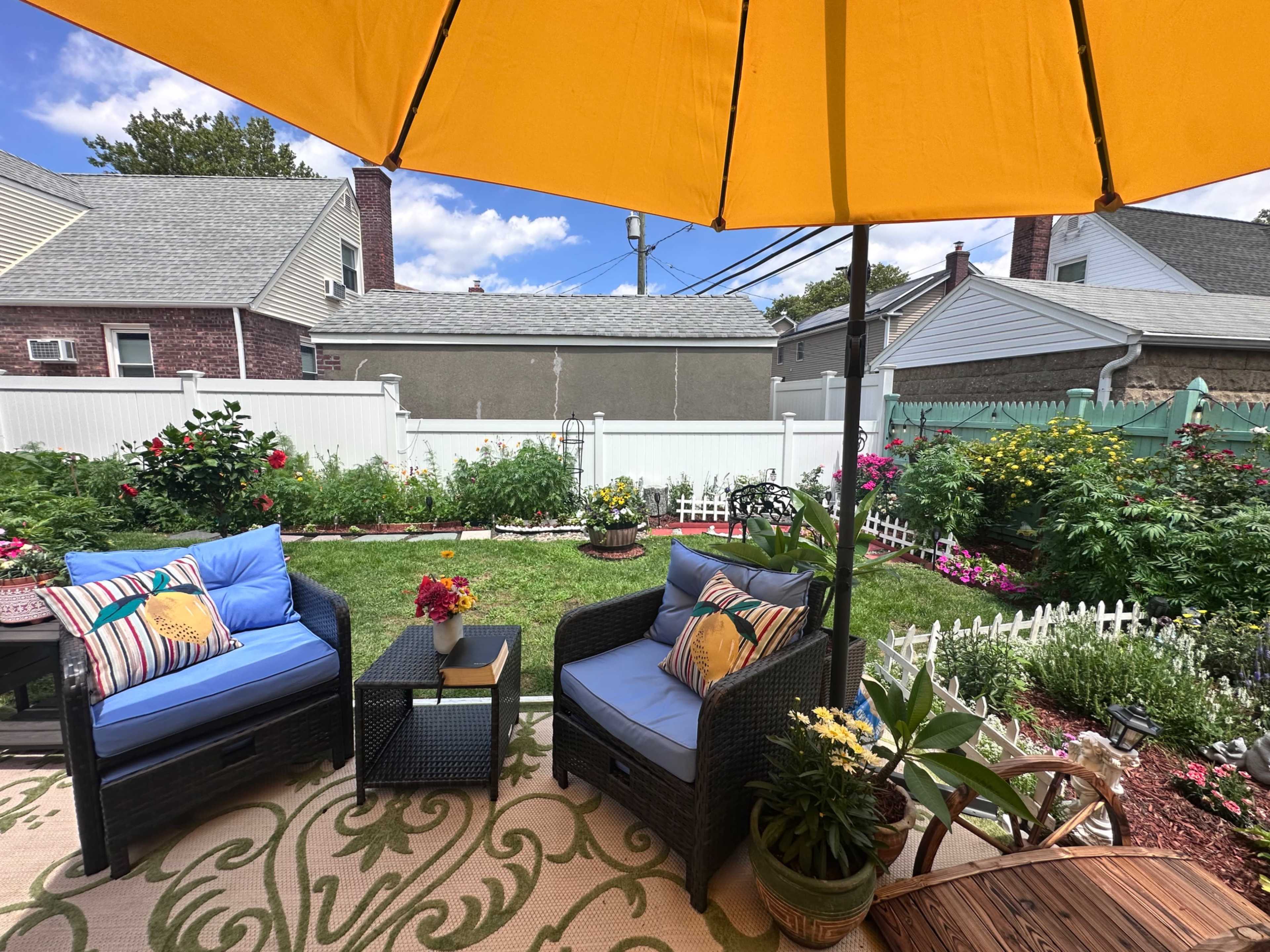 The image shows a backyard patio with two blue chairs, a small table, and a large yellow umbrella, surrounded by vibrant flower beds and a white picket fence.