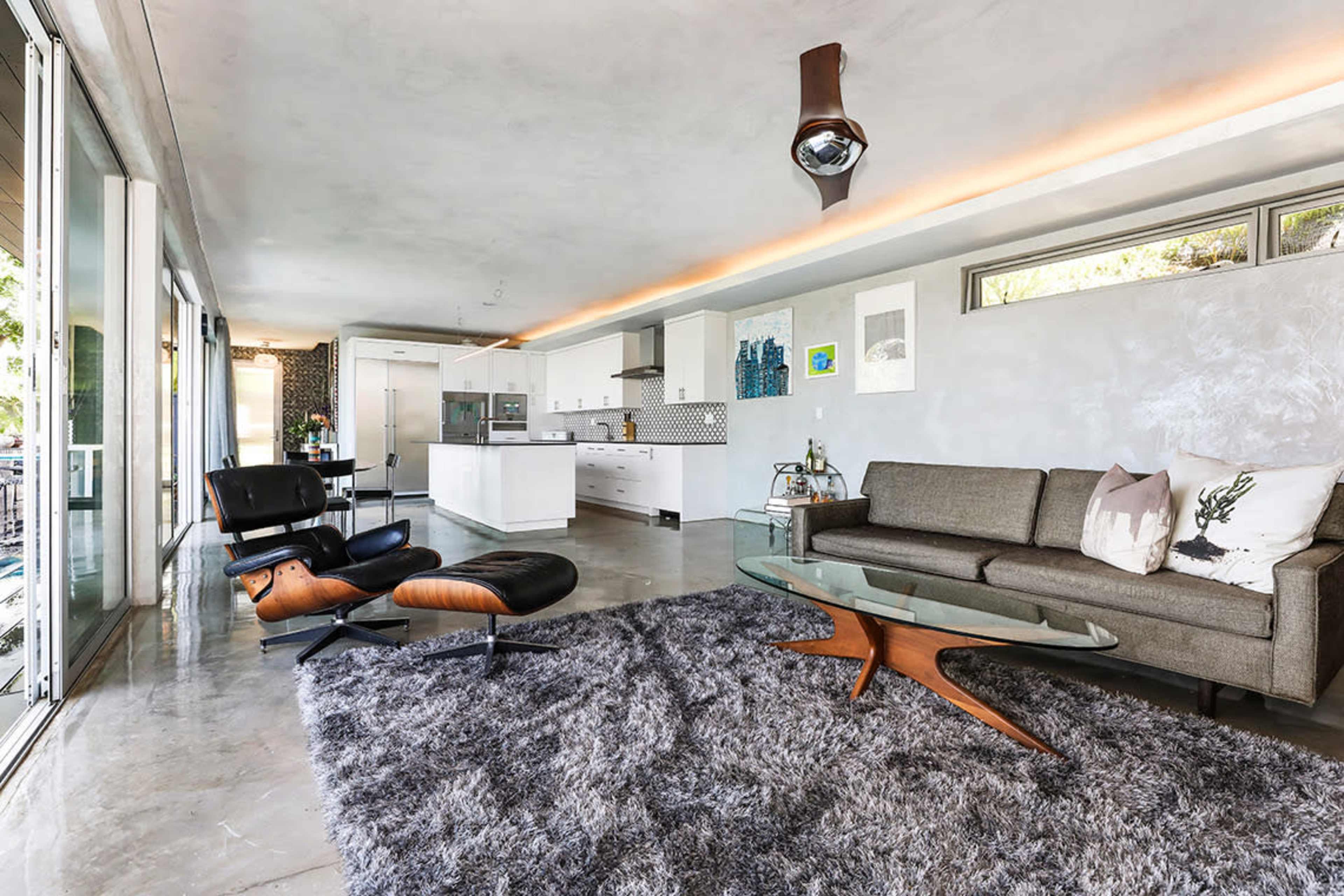 The scene shows a modern living room with a sleek sofa, an angular coffee table, a rug, and a kitchen area visible in the background.