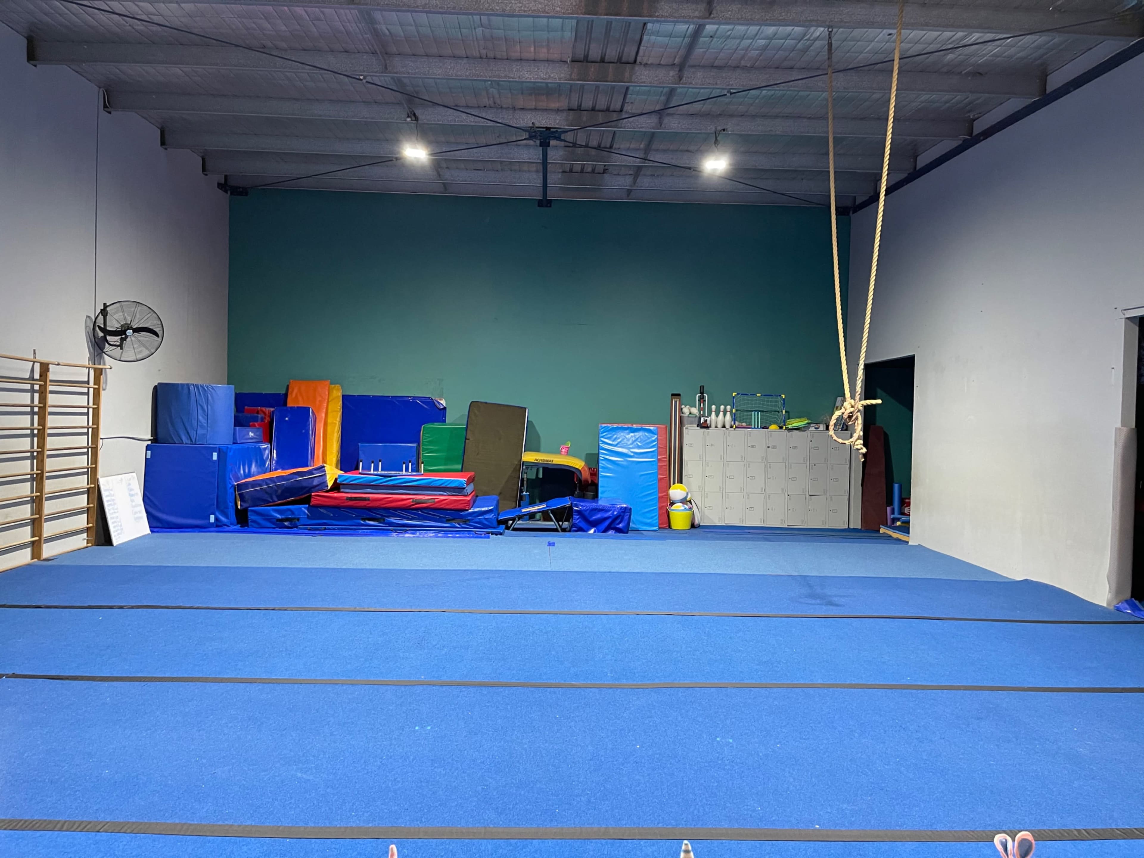 A gymnasium floor covered in blue mats, surrounded by various gym equipment, including colorful mats and a climbing rope.