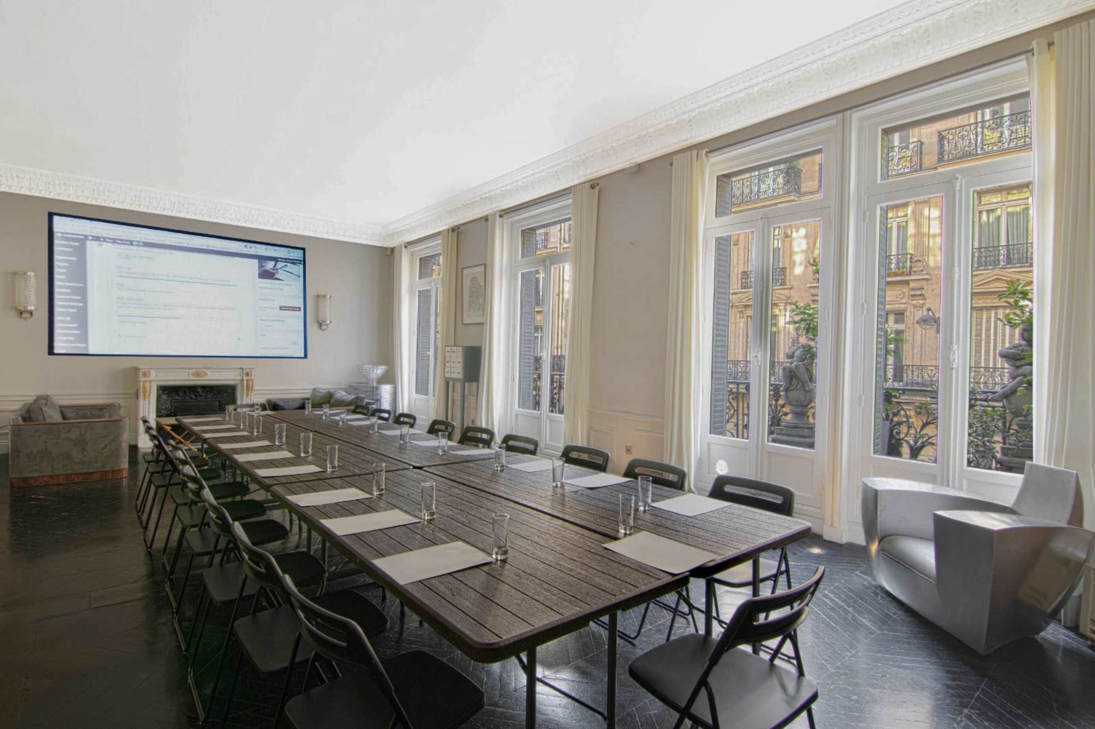 The image shows a modern conference room with a large table set for a meeting, flanked by windows with views of city architecture.