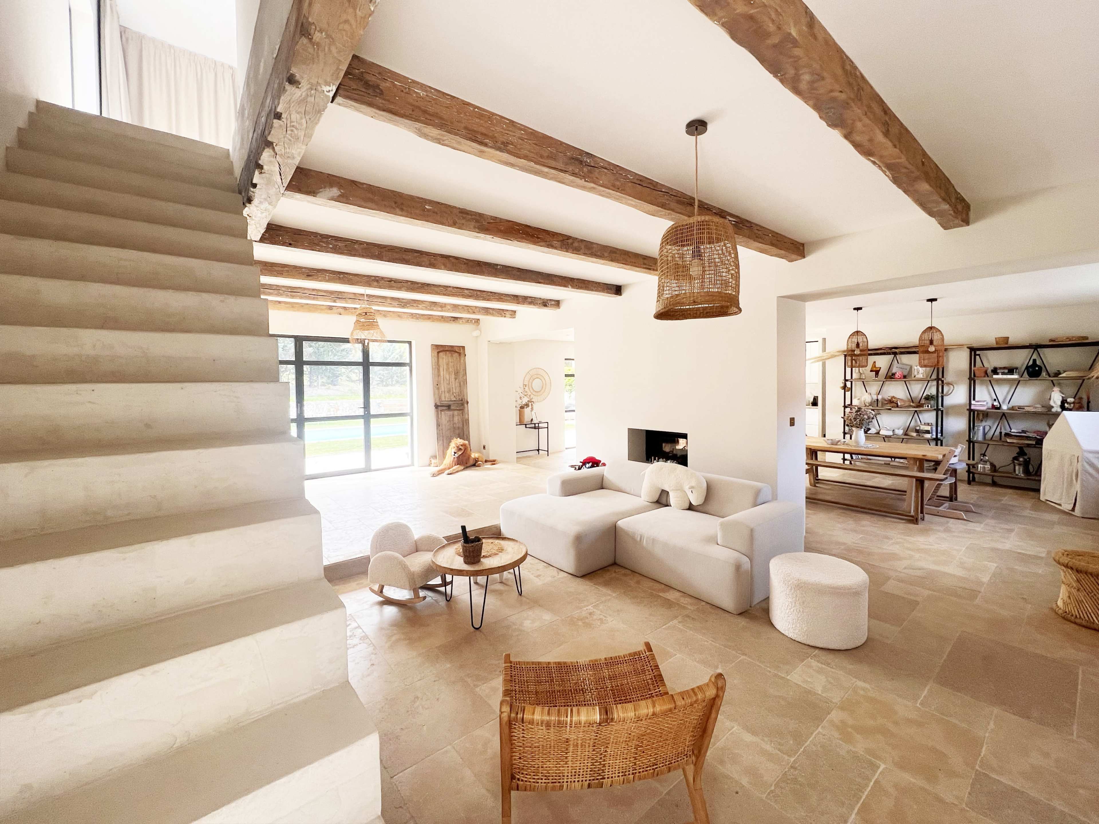 The image showcases a spacious living area with a staircase, exposed wooden beams, comfortable seating, and a large window allowing natural light to fill the room.