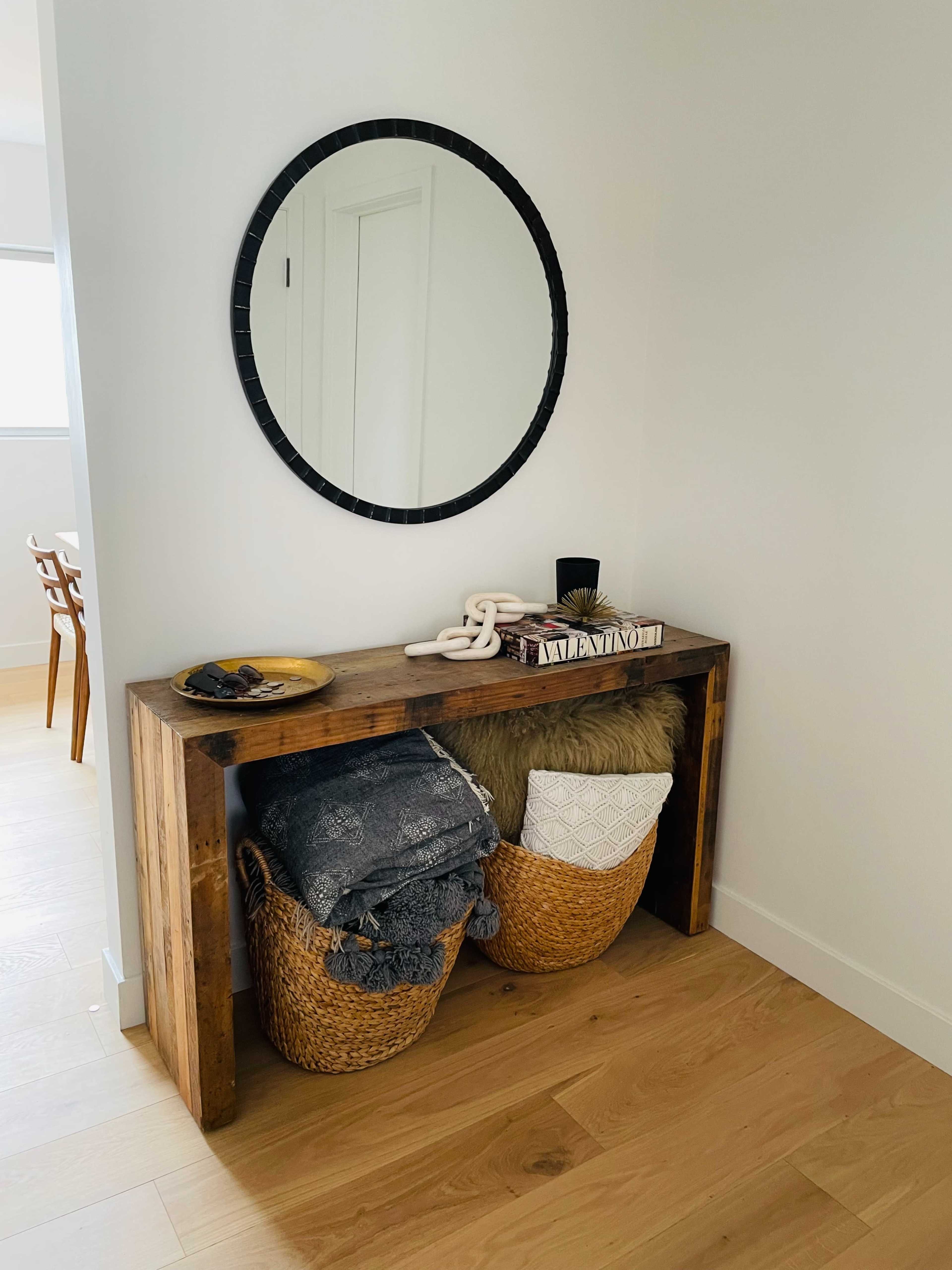 A wooden console table is positioned against a wall, reflecting a round mirror, with two woven baskets filled with blankets and pillows underneath.