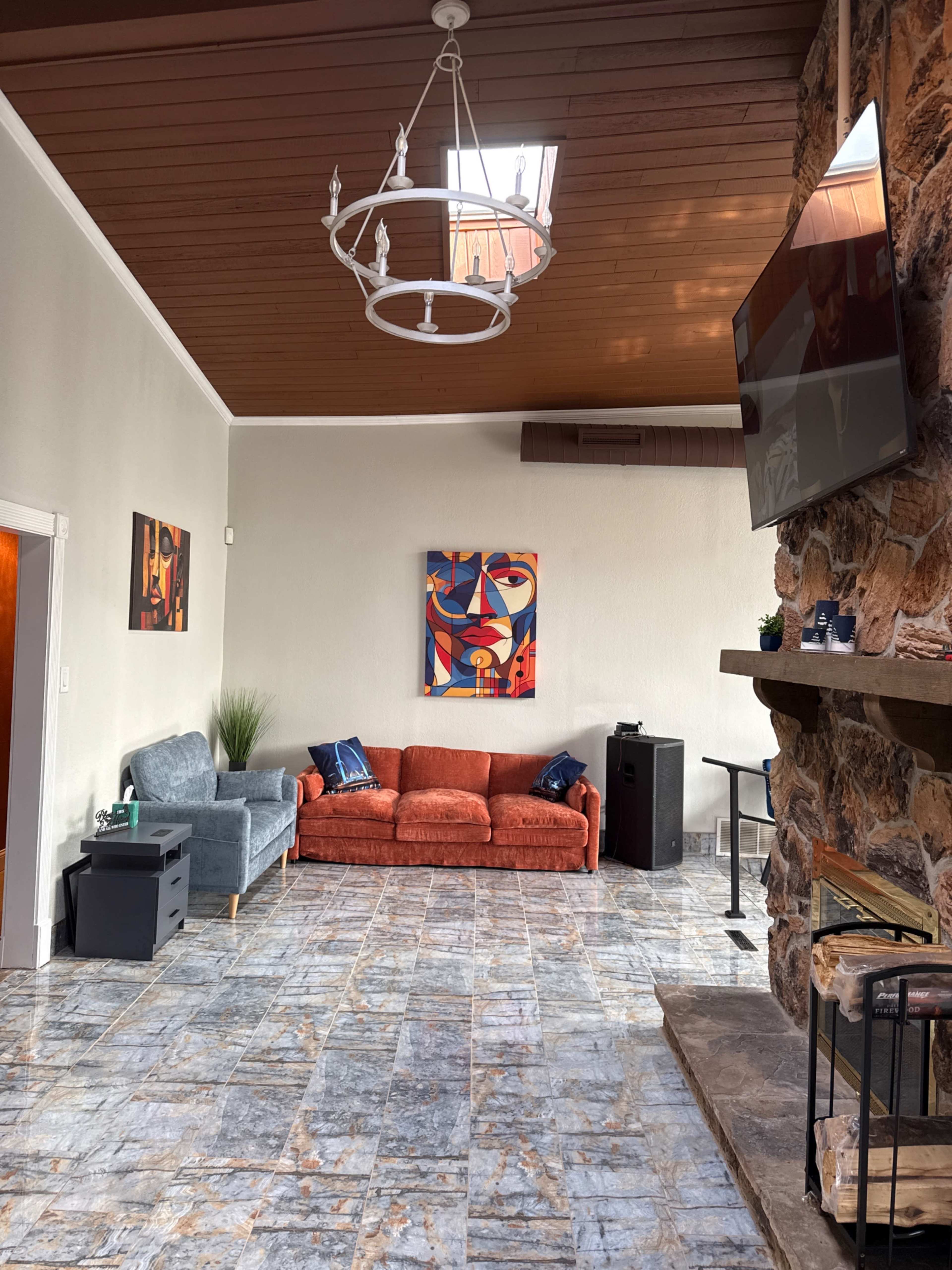 The image shows a spacious living area featuring a red couch, a blue armchair, a stone fireplace, and a mounted television, with a piece of abstract art on the wall.