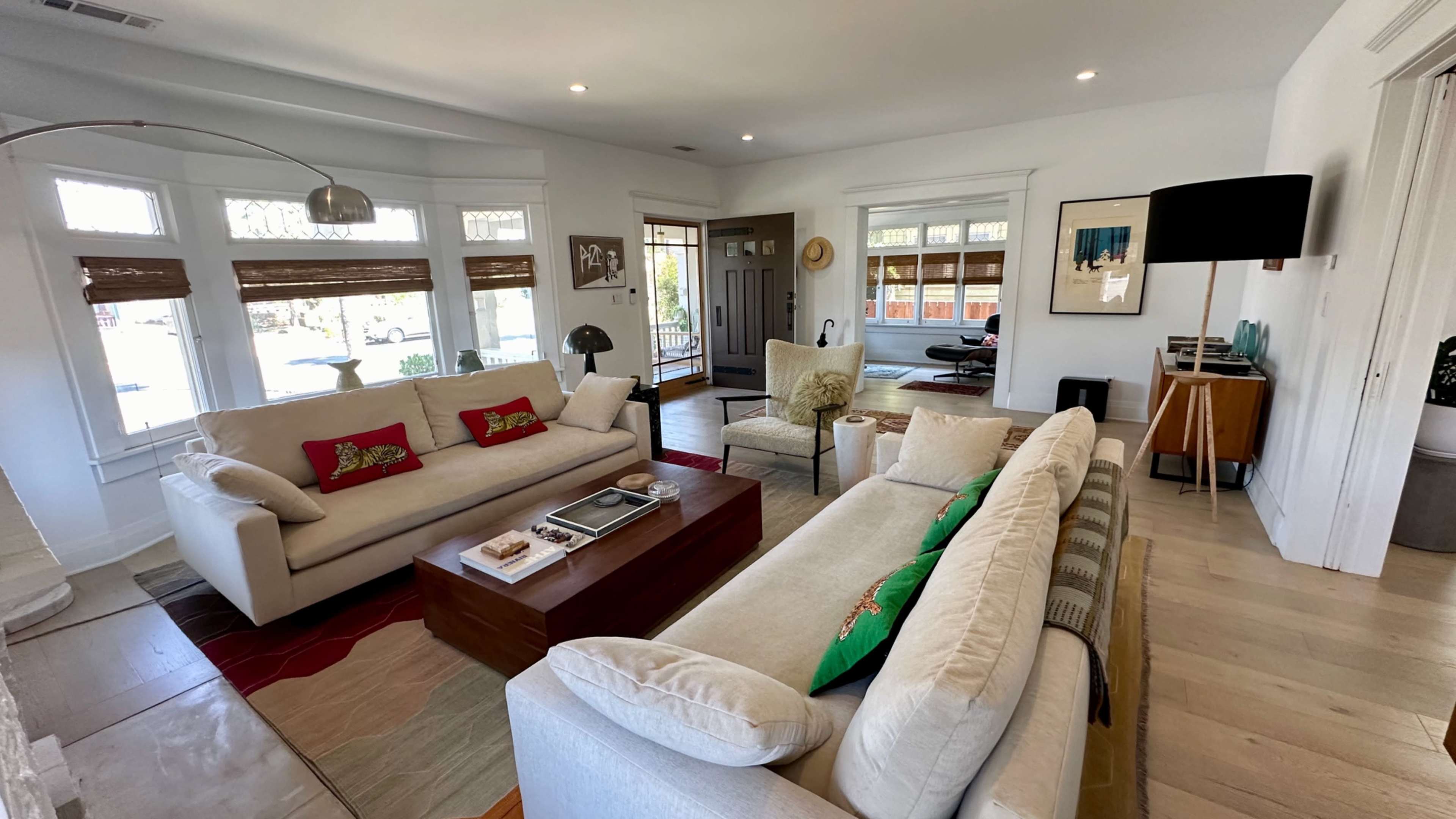 The image shows a bright and spacious living room with two sofas, a wooden coffee table, and a mix of decor items, including a floor lamp and various artworks.