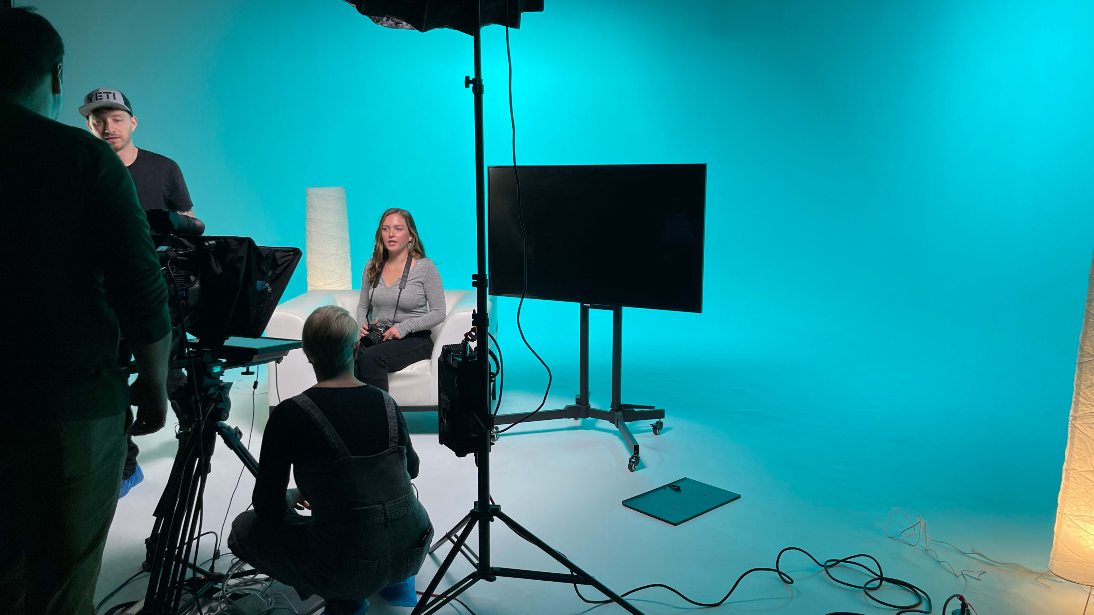 A woman sits on a sofa in front of a large screen, while two crew members prepare equipment on a brightly lit set with a blue backdrop.