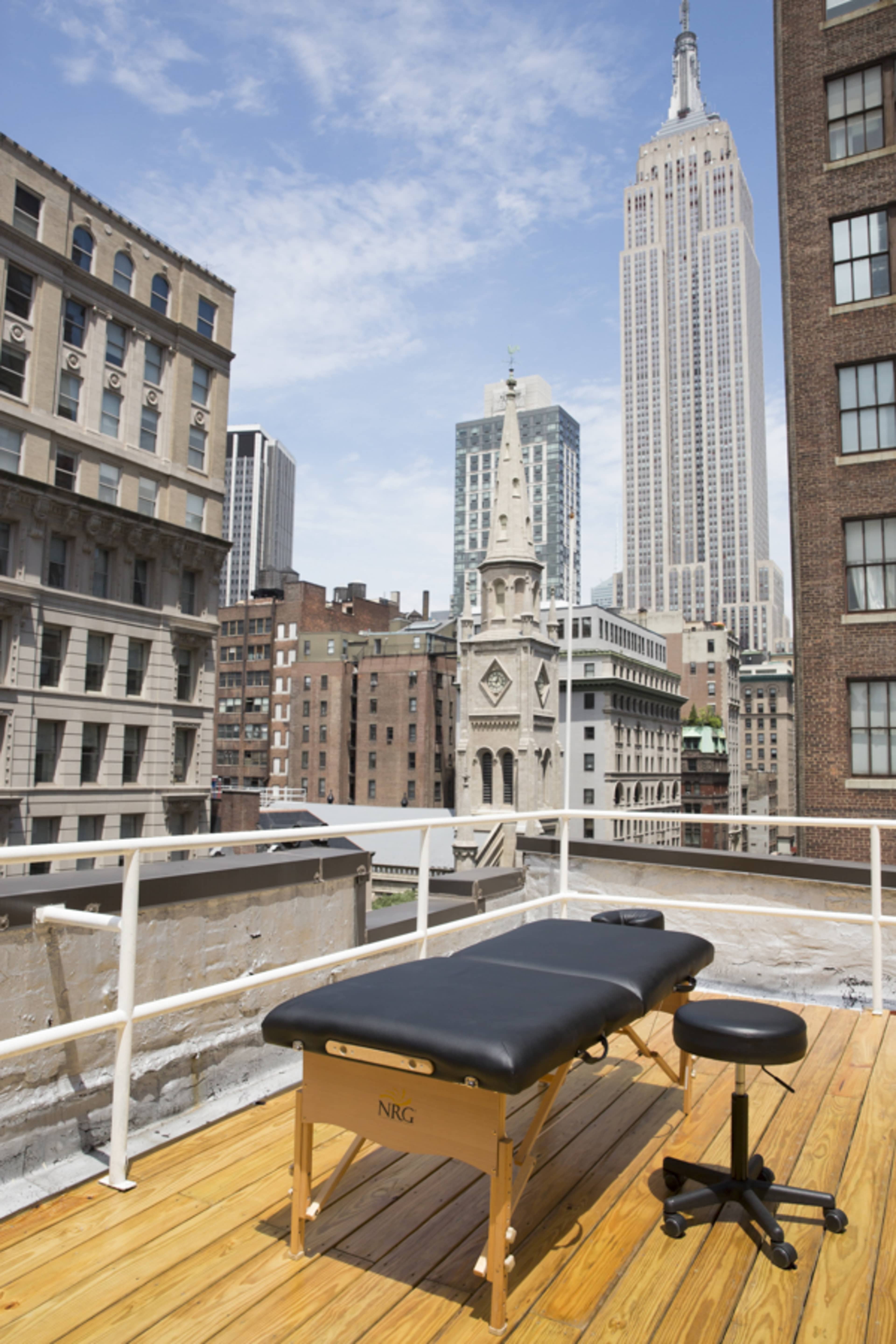 A massage table and stool are positioned on a rooftop deck with the Empire State Building visible in the background.