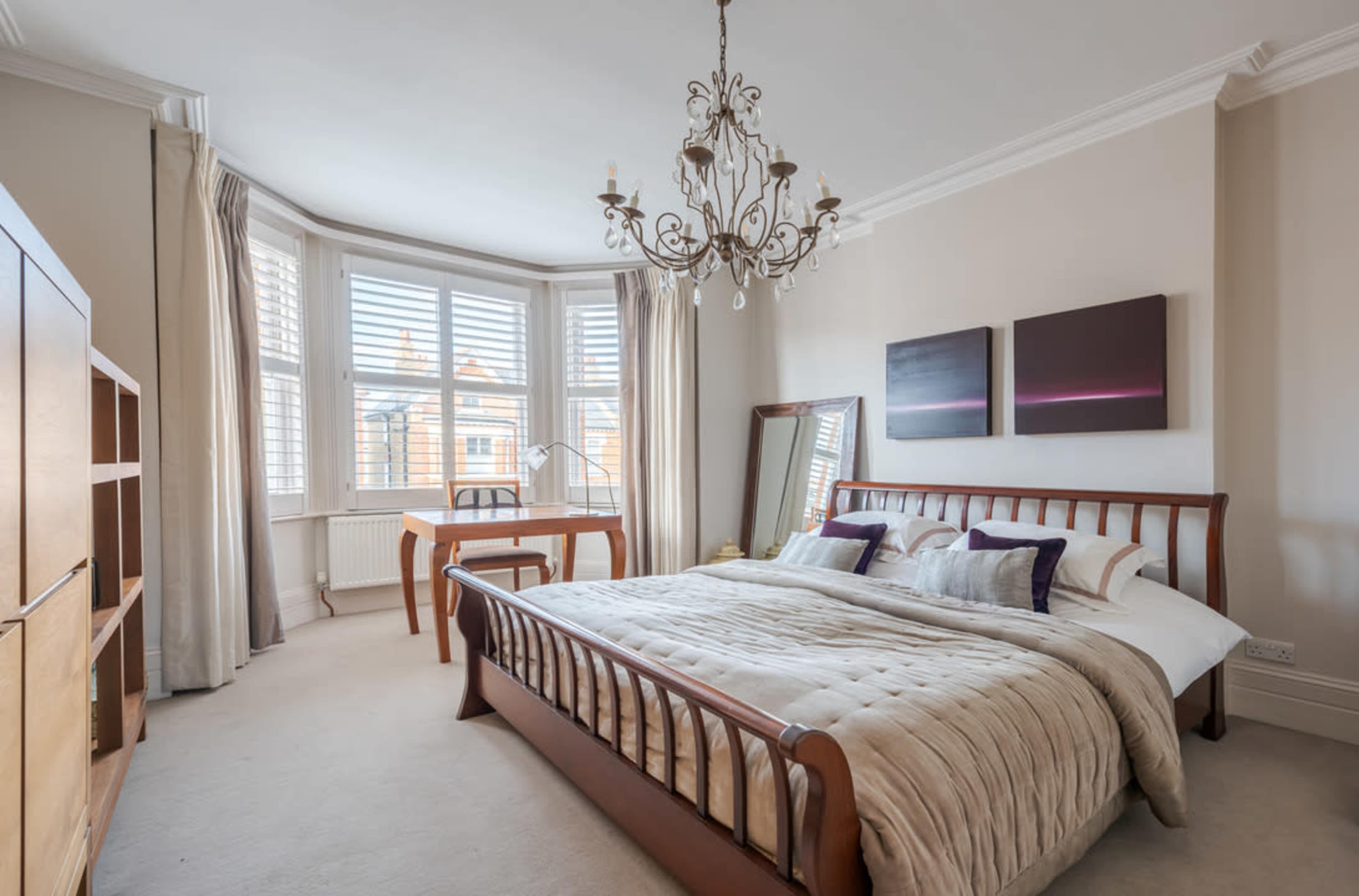 The image shows a spacious bedroom with a large window, a wooden bed with a beige quilt, a chandelier, a wooden bookshelf, and a desk.
