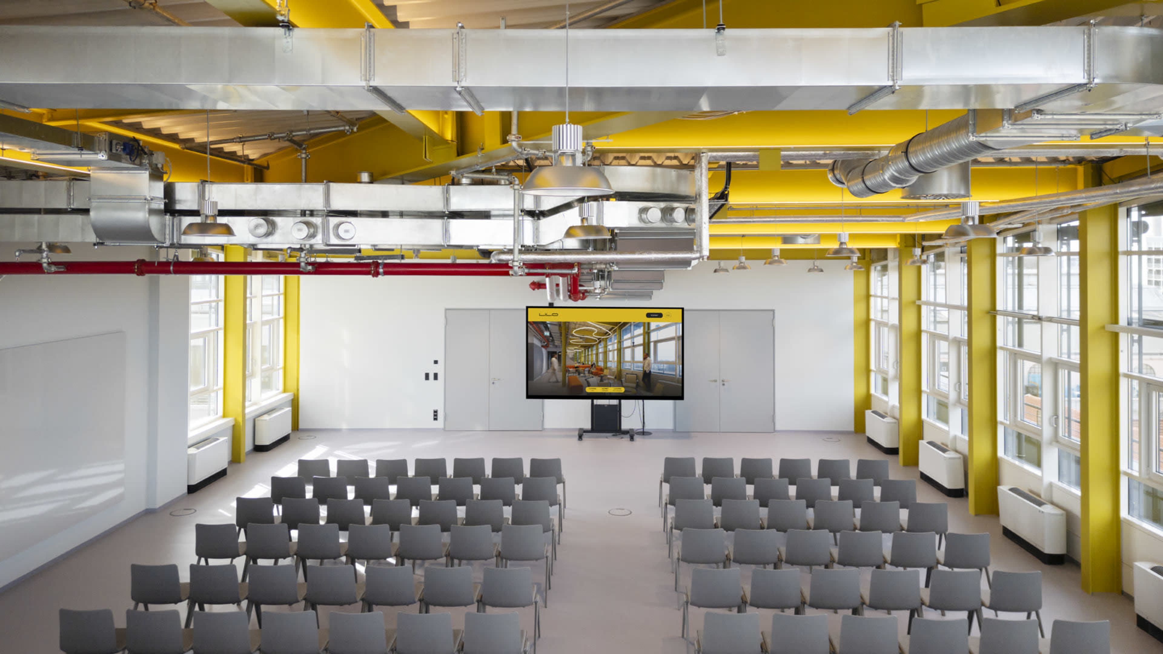The image shows a modern classroom or conference room with rows of gray chairs facing a large screen, illuminated by bright yellow beams and large windows.