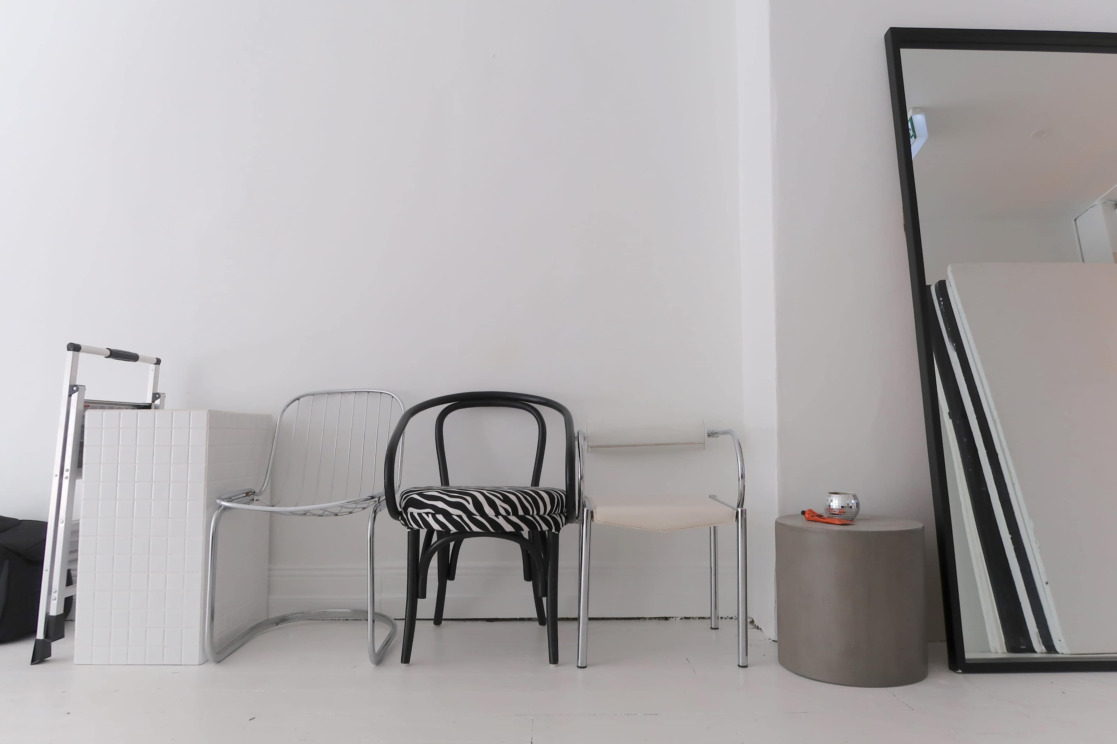 The image shows a minimalist interior featuring a gray metal table and three diverse chairs, including a striped chair, against a white wall with a large mirror nearby.